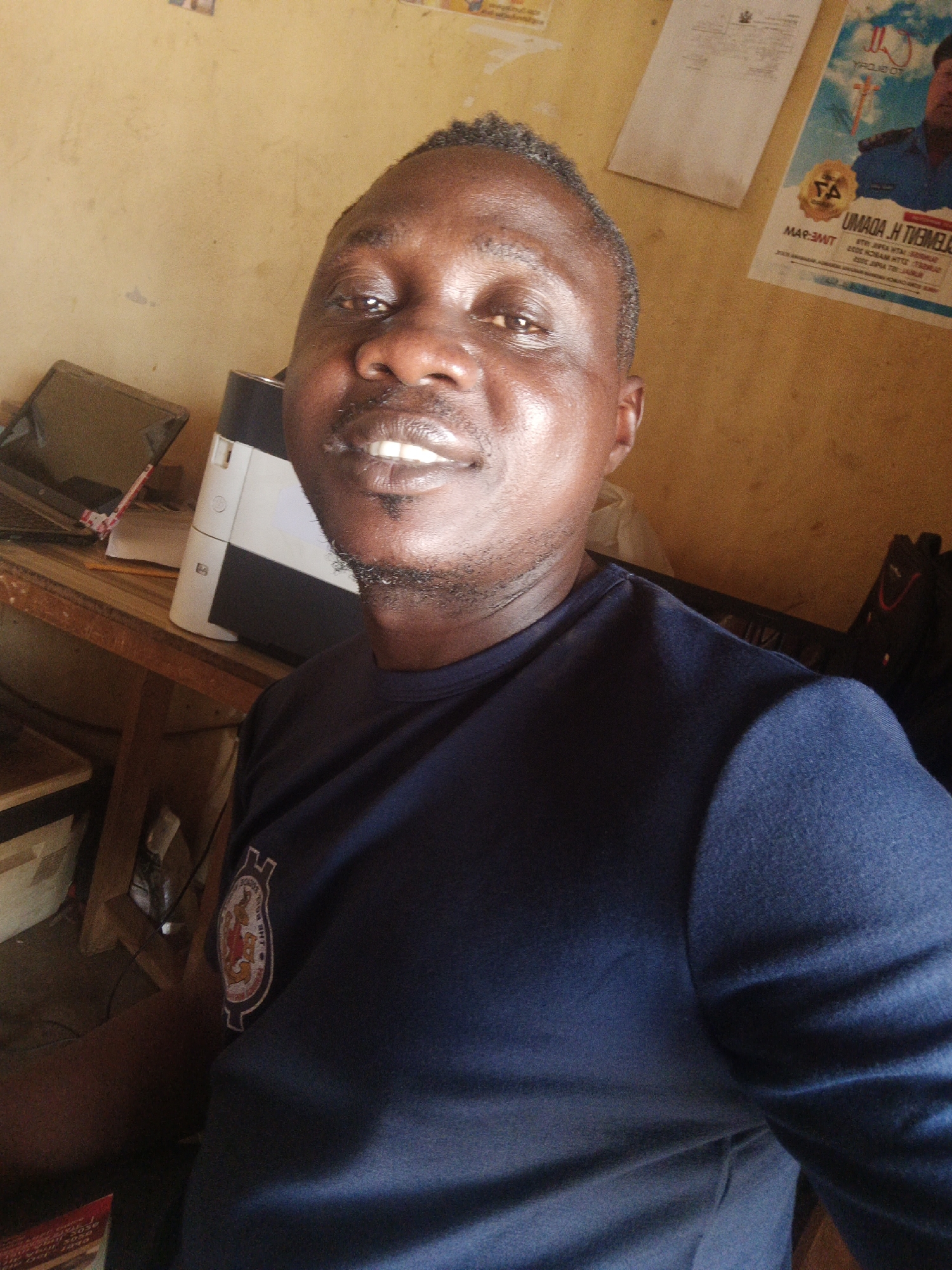 The image is a selfie of an adult man taken indoors in Akwanga, Nigeria. The man, who appears to be of African descent, has dark skin, short dark hair, and a neatly trimmed goatee. He is smiling broadly, showing his white teeth, and looks directly at the camera with a happy and confident expression. He is wearing a dark blue short-sleeved t-shirt with a circular crest or logo on the left chest. This emblem features text, partially visible, which reads "LAGOS REGIONAL ACADEMY" and "THE ROYAL" surrounding a central design.
The background shows a light brown or tan wall. To the man's upper right, a poster is visible, featuring an image of another man and text that includes "MAP-3MT" and "UMADA. H. TREMELOH," along with an unreadable emblem and smaller lines of text. Another white document is also affixed to the wall. To the man's left, a wooden desk holds a closed dark-colored laptop, a white and black printer, and a pencil. A black bag or backpack is partially visible on the right side of the background. The lighting is bright, suggesting daytime, and illuminates the man's face. The setting appears to be a casual office or workspace.