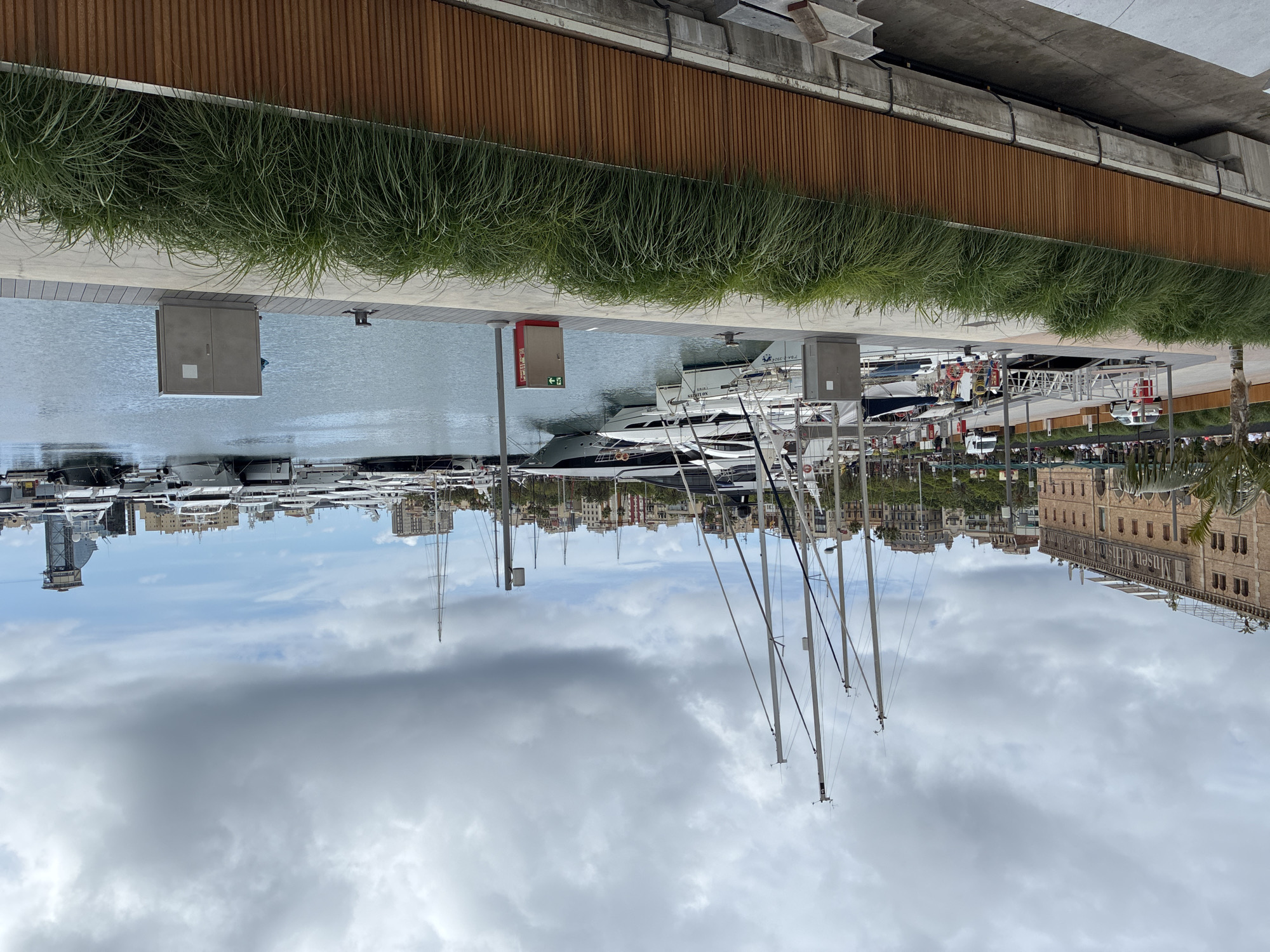This image captures a harbor in Barcelona, Spain. It depicts several yachts and boats docked in the water. The backdrop includes the city skyline, with a prominent building identified as the "Museu d'Història de Catalunya." The sky is overcast with clouds. A section of green grass and a wooden structure appear in the foreground along the harbor's edge.