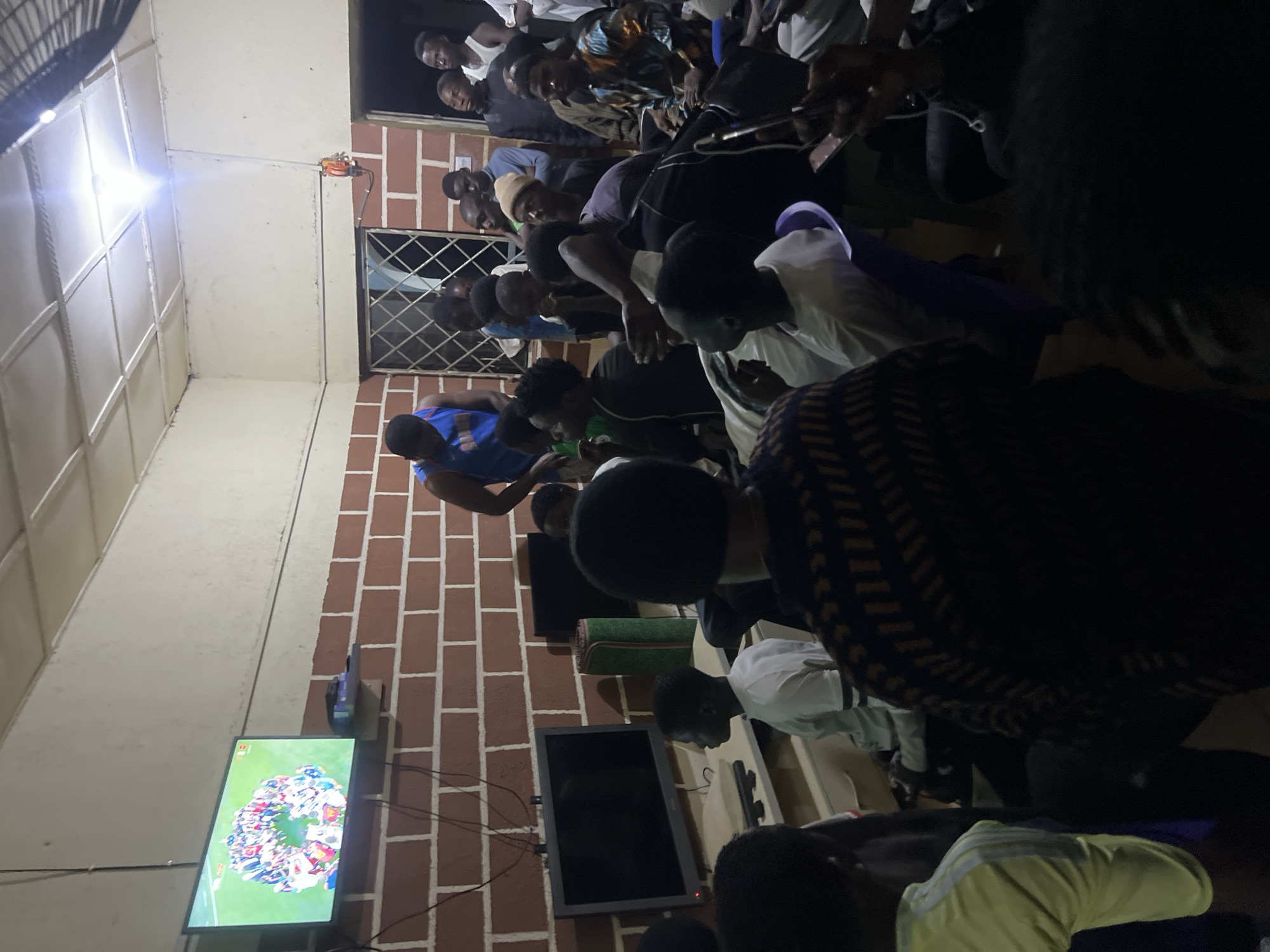 The image depicts an indoor gathering of approximately 20 young men and boys, likely in Jos, Nigeria, watching a television screen displaying a sports event. The room appears to be a communal space, possibly a living room or recreational area, with exposed brick walls and a tiled ceiling. The lighting is dim, with a bright overhead light illuminating the ceiling.
Several individuals are seated on chairs and benches, while others stand, all facing the television. The individuals are predominantly dressed in casual clothing, including t-shirts, jerseys, and shirts. One person in the foreground is wearing a distinctive patterned shirt. The atmosphere suggests a shared viewing experience, with some individuals engaged in conversation or observation.
Objects in the scene include the television, which is mounted on the wall, along with two smaller black monitors below it. Various electronic devices and cables are visible. A green cylindrical object, possibly a mat or rug, is placed near the monitors. The overall impression is one of a casual social gathering focused on a shared activity.