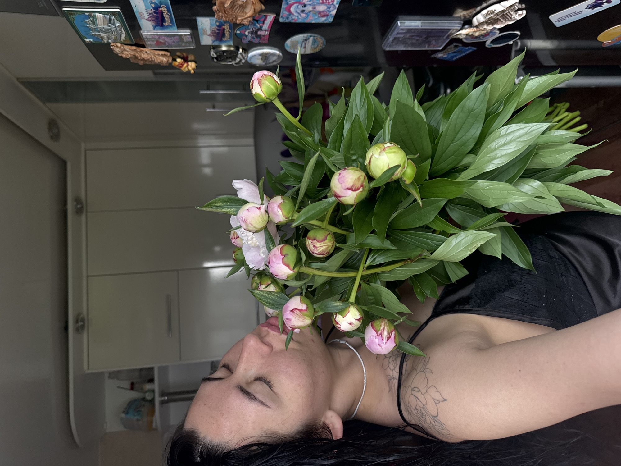 In a domestic kitchen setting, a woman with dark, wet hair is embracing a large bouquet of pink and white peonies. Her eyes are closed, and she is leaning in to smell the flowers, her lips slightly parted. She is wearing a black silk camisole with thin straps. A delicate beaded necklace adorns her neck, and a floral tattoo is visible on her left shoulder and upper arm.
The kitchen features white cabinetry and a stainless-steel refrigerator adorned with various magnets and souvenirs. The background suggests a typical apartment kitchen, common in cities like Chaikovsky, Russia, known for its industrial and residential areas. The lighting is soft, indicating it might be daytime, possibly overcast, given the diffuse light in the room. The overall scene conveys a moment of quiet enjoyment and appreciation for nature's beauty within a home environment.