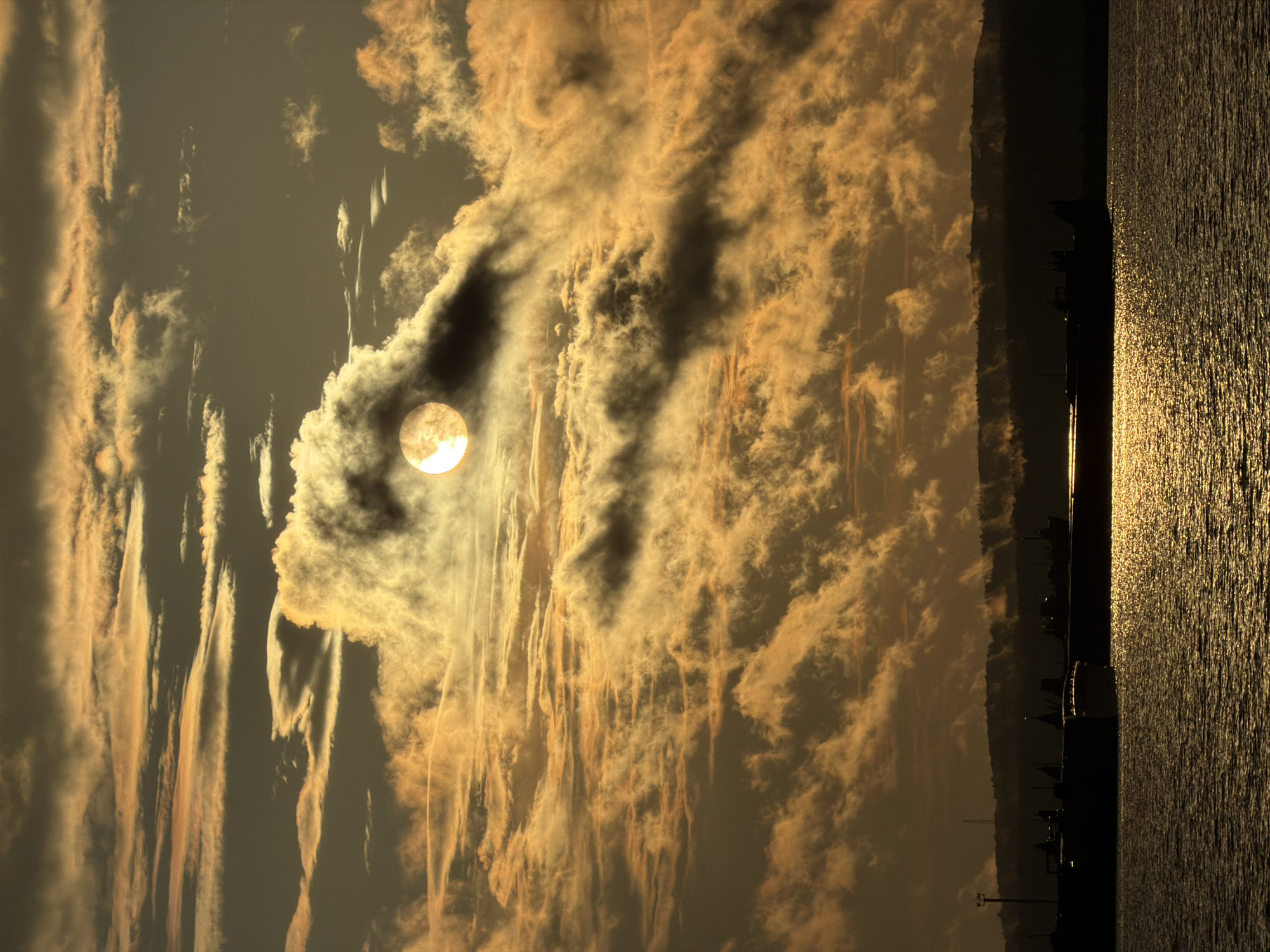 A sunset is captured over a body of water in Chaikovsky, Russia, with the sun partially obscured by dense, golden clouds. The sun's rays create a shimmering reflection on the water's surface. In the foreground, several silhouetted barges and ships are docked along a shoreline, with a dark, tree-lined horizon visible in the distance.