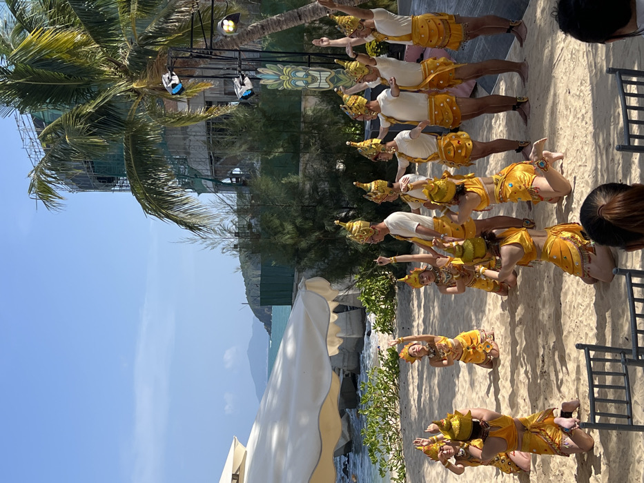 A group of performers in yellow and white traditional attire are on a beach. They wear elaborate headdresses. Their costumes feature ornate details and unique accents. The background includes a beach with palm trees, a partially constructed building, the sea, and the sky. Chairs are placed on the side and near the performers. The overall scene showcases a cultural performance by the beach.