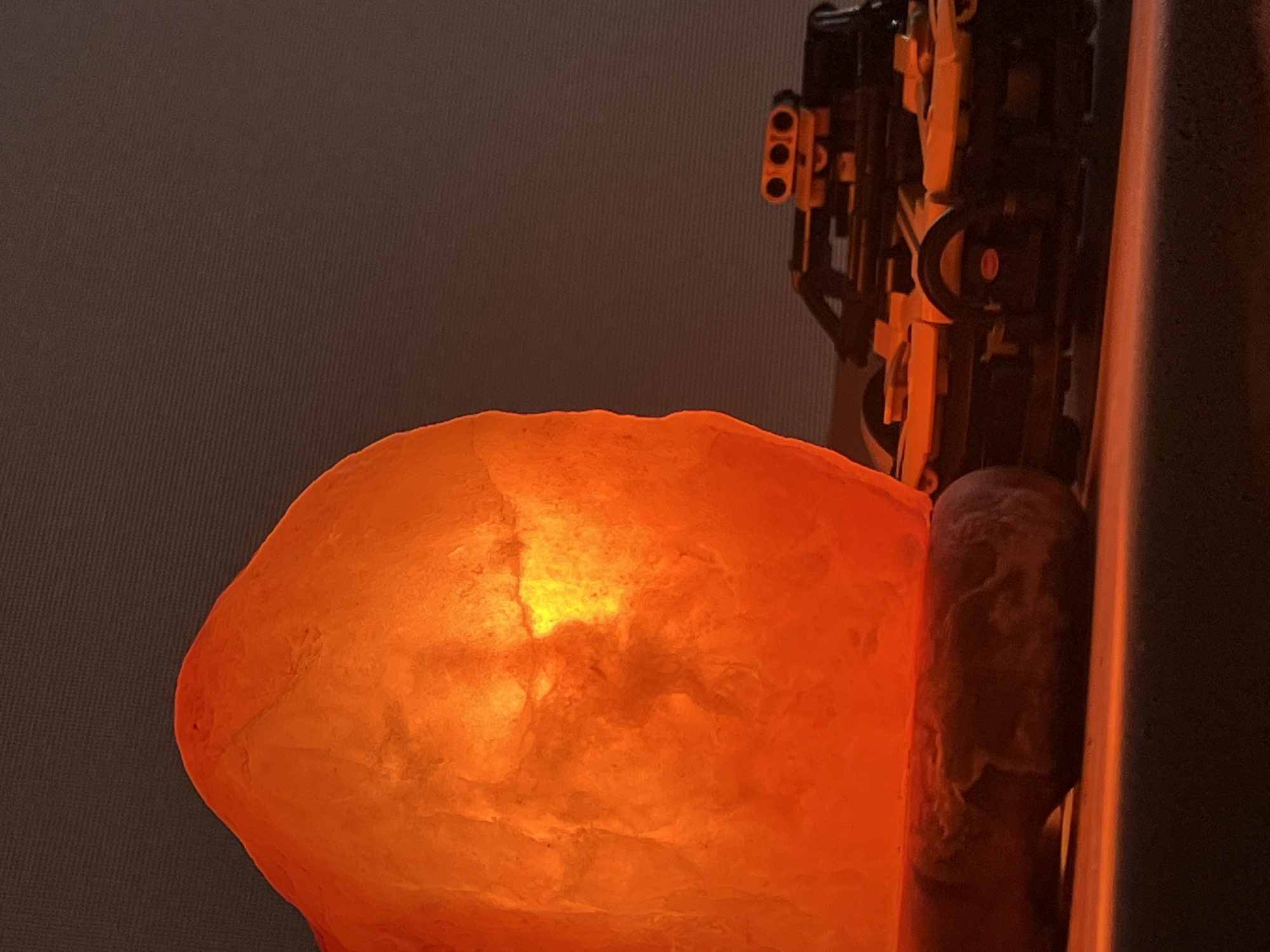 The image captures a still life composition featuring an illuminated object and a constructed model in a dimly lit environment. A large, irregularly shaped Himalayan salt lamp is the central focus, glowing with an intense, warm orange and yellow light. Its rough, crystalline surface shows variations in translucence, with brighter areas indicating the internal light source.
To the upper right of the salt lamp, a complex structure built from black and yellow/orange LEGO bricks is partially visible. This construction features several distinct components, including a section with three small, circular openings, suggesting a detailed model or device. Below this LEGO structure and to the right of the salt lamp, a dark, textured, cylindrical or pillar-like object is present.
The background is a uniform dark grayish-brown, showing a subtle diagonal striped or woven texture. The overall lighting is low, with the salt lamp being the primary source of illumination, casting a warm glow on itself and subtly highlighting parts of the other objects. The absence of strong ambient light suggests the image was taken in a darkened room, possibly during the evening or night.
There are no people, explicit activities, or discernible text in the image. The composition highlights the contrast between the organic, glowing salt rock and the intricate, manufactured LEGO structure.