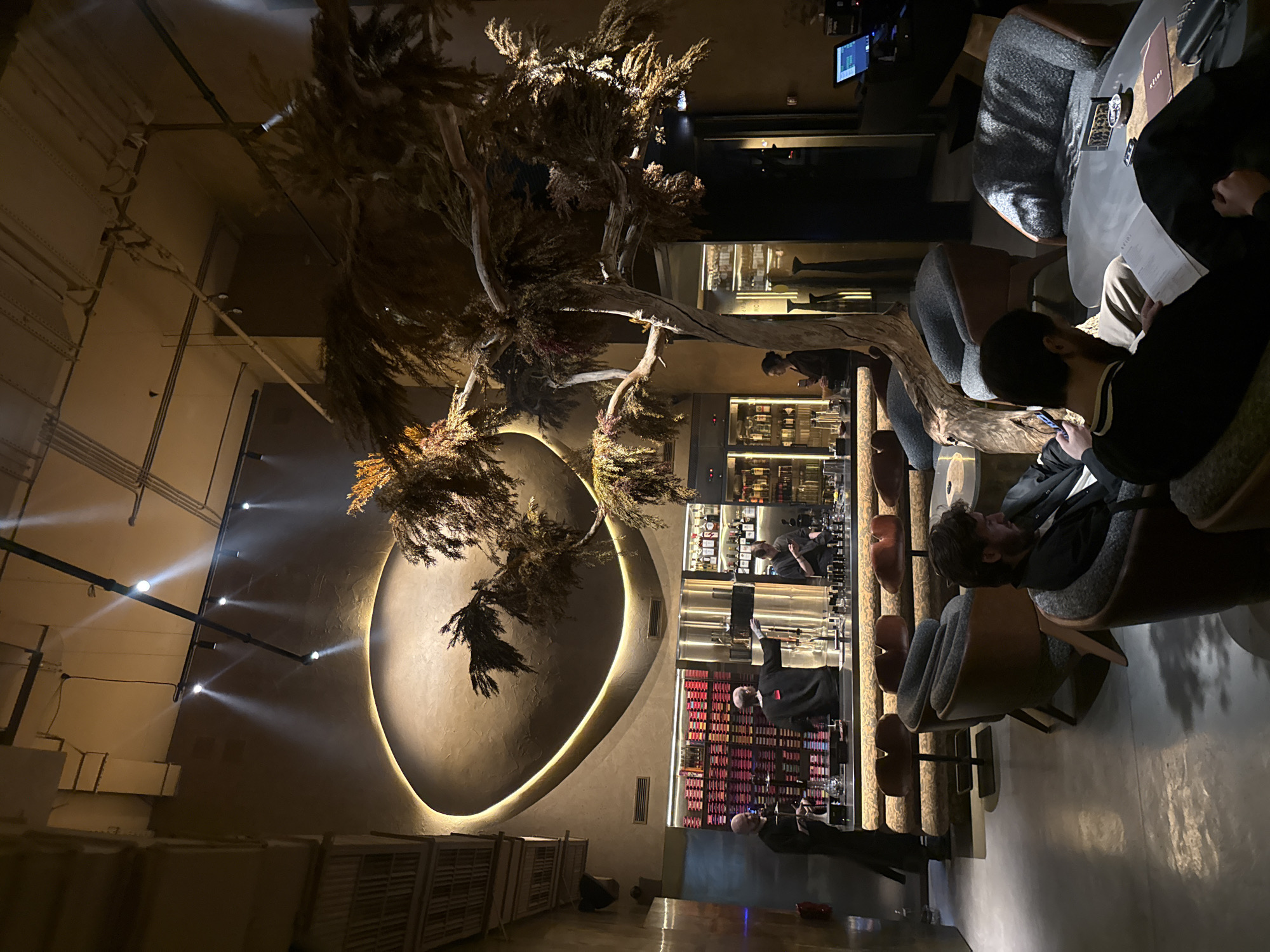 The image depicts an interior scene, likely a bar or lounge, in what is stated to be Khimki, Russia. The scene is dominated by a large, decorative tree structure that seems to be artificial or dried, placed centrally in the space. The lighting is dim and ambient, with a prominent circular or oval illuminated backdrop against a beige wall. A bar area is visible in the background, with shelves of liquor bottles and bartenders at work. Several people are seated at tables or at the bar, engaged in conversations or using their phones. Seating appears to be plush and modern, with a mix of chairs and bar stools. In the foreground, people are seated around a round table, some are looking at menus or using electronic devices. The floors are shiny, suggesting they are polished concrete or a similar material. The overall setting conveys a sense of upscale urban sophistication.