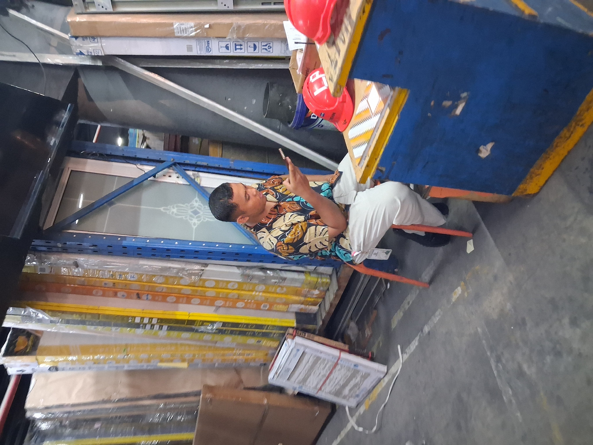 A man, dressed in a short-sleeved, floral-patterned shirt and light-colored pants, sits on an orange stool in what appears to be a warehouse or storage facility in Depok, Indonesia. He is focused on his mobile phone, holding it in his right hand. The man is positioned in front of a blue and yellow counter, on which two red hard hats are visible. Behind him, shelves are stacked with various items, including what seem to be construction materials or building supplies. The floor is concrete, with some yellow and white markings. The overall environment suggests a commercial or industrial setting.