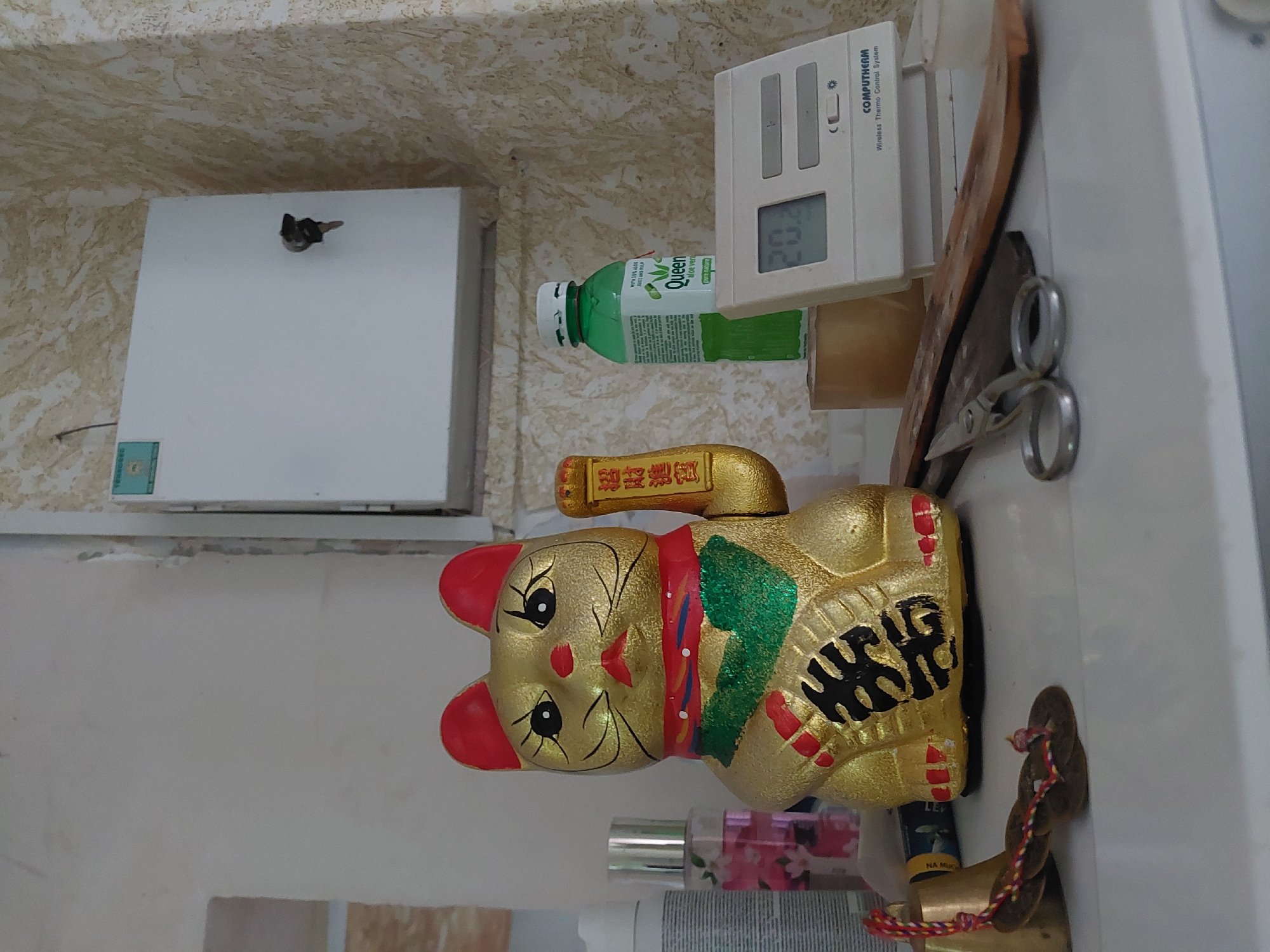 An indoor scene, rotated 90 degrees clockwise, displays a collection of household items on a white surface, likely a shelf or countertop, against a light-colored, textured wall in Orhei, Moldova.
The most prominent object is a golden Maneki-neko (lucky cat) figure, lying on its side. It has red ears and nose, a red and green collar-like scarf, and black stylized Chinese characters on its body and on a gold ingot it holds. The characters visible on the ingot are "招財進寶" (Zhāo Cái Jìn Bǎo), meaning "ushering in wealth and prosperity." To the right of the cat is a traditional Chinese-style coin or charm with a red, yellow, and blue braided string attached. Below the cat, a clear spray bottle with a pink floral pattern is partially visible.
Above the cat, positioned on the white surface, are a green bottle and a white electronic device. The green bottle, labeled "Queen aloe vera pure nature," contains a light green liquid. Next to it is a "COMPUTHERM Wireless Thermo Control System" device, displaying "20.3" on its screen. Behind this device are a pair of silver-colored scissors and two pieces of wood, one a thin plank and the other a darker, thicker block.
To the far left of the image, mounted on the wall, is a white rectangular box or cabinet with a set of keys hanging from a hook on its top edge. The wall behind these objects is a light cream or beige color with an uneven, slightly textured pattern. No people or activities are depicted.