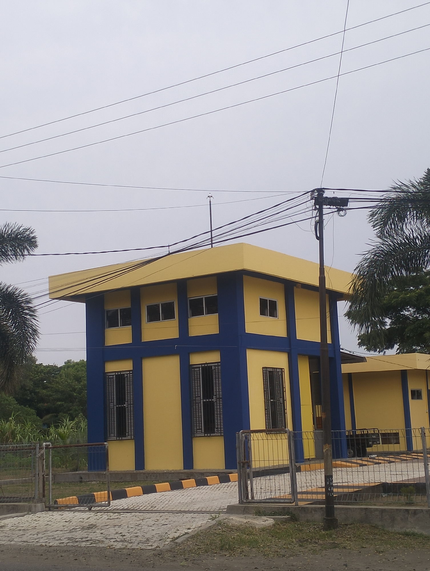 A two-story building with a yellow facade and blue accents is visible under an overcast sky. The building has multiple windows with dark grates. A street with a yellow and black striped curb runs in front of the building, bordered by a metal fence. Several power lines are strung across the sky above the building and a palm tree is visible on the right side. The scene is set in the city of Lhokseumawe, Indonesia.