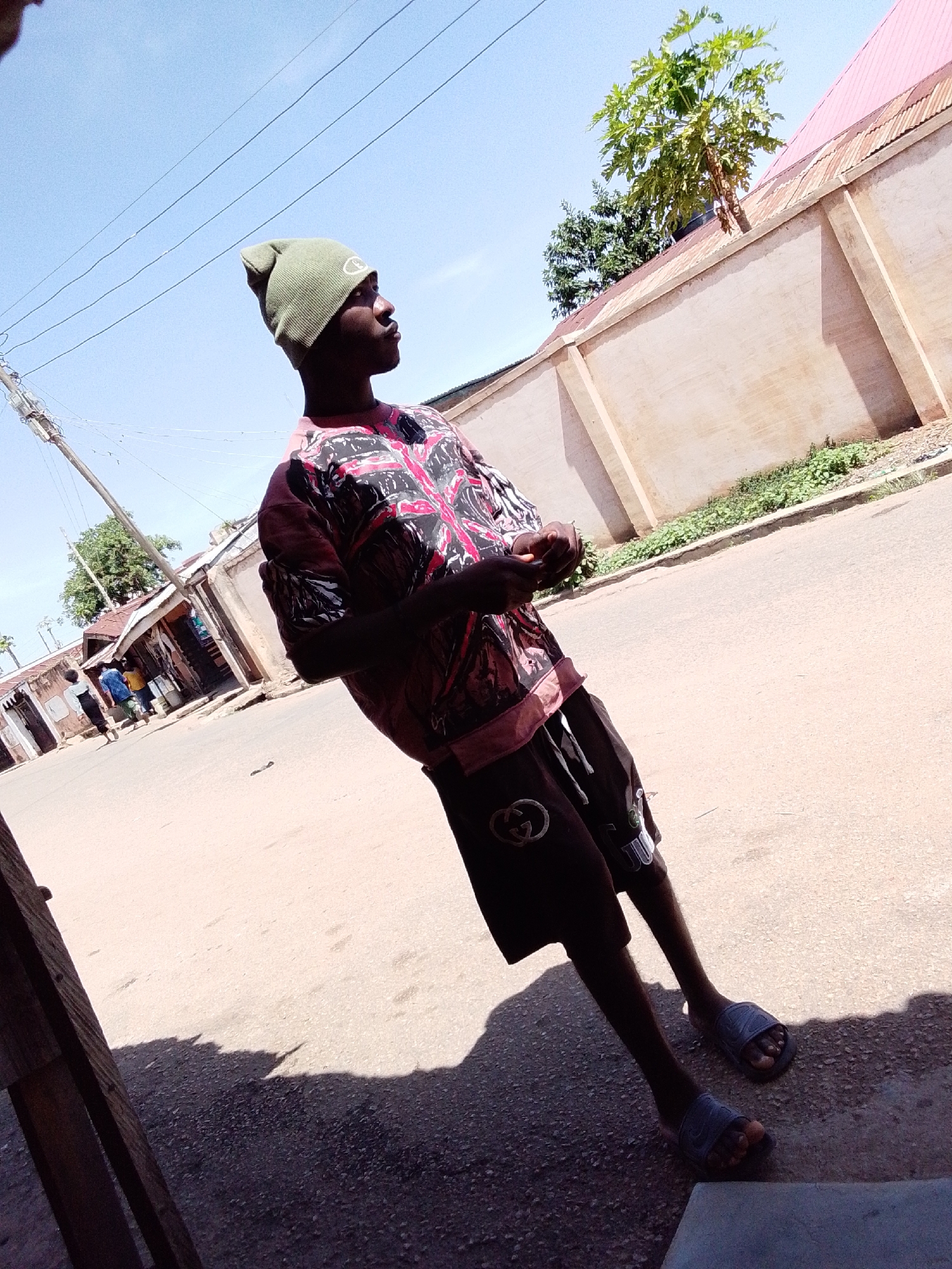 A young man stands on a paved road under a clear, bright blue sky in Jos, Nigeria, captured from a low angle. He is positioned slightly off-center to the right, looking towards the right side of the frame. He wears a dark-colored t-shirt with a striking red, black, and white graphic pattern, dark shorts featuring a white "Gucci" logo, a light green beanie with a circular "CR" logo, and grey slide sandals. His hands are clasped loosely in front of him.
The setting appears to be a residential or commercial street. To the left, a utility pole stands, with multiple wires stretching across the sky. In the background, several single-story buildings line the street, some with simple storefronts, and a few individuals are visible walking further down the road. To the right, a tall, light-colored wall with a pink/red corrugated metal roof runs alongside the road, behind which a tree with large green leaves (possibly papaya) and other vegetation are visible. Strong shadows cast on the ground indicate a sunny day. The road surface is a mix of asphalt and dirt.