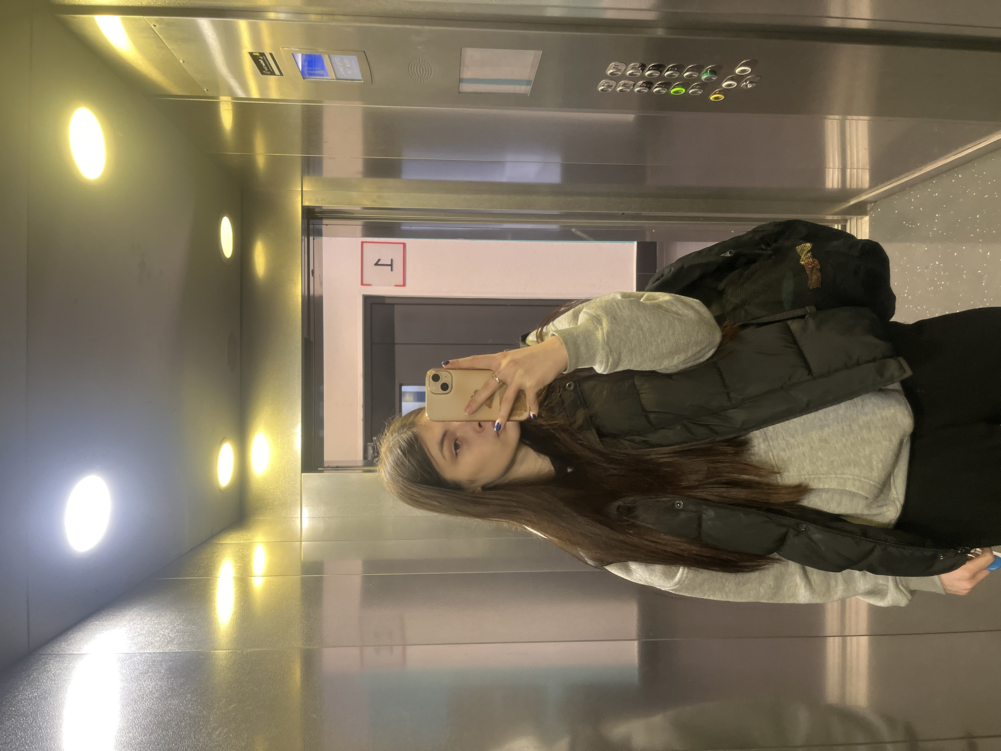 A woman is taking a selfie in an elevator. She is wearing a gray hoodie, a black puffer vest, and has a black backpack on her back. Her hair is long and brown. The elevator has stainless steel walls and ceiling. There are round, recessed lights on one wall. The elevator floor indicator shows the number "1" in a red box. The elevator control panel is visible above the woman's head. The location is unknown; Saint Petersburg, Russia is not discernible from the image.