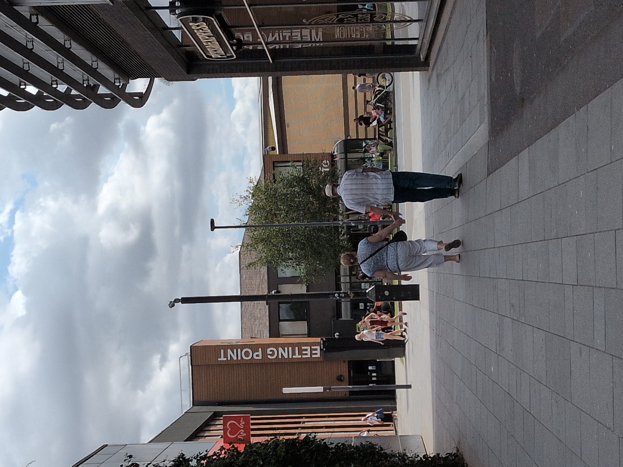The image captures a street scene in Telford, United Kingdom, likely during daytime, with a slightly overcast sky. In the foreground, a couple walks hand-in-hand away from the camera, the man wearing a striped shirt and dark trousers, and the woman in a floral top and light-colored trousers. To the left, a building with a vertical sign reading "MEETING POINT" is visible. Further to the right, the facade of a "Pizza Express" restaurant is prominent, featuring its distinctive sign. Other individuals and groups are scattered throughout the scene, including people walking, sitting at tables, and standing near shops. The environment appears to be a pedestrianized area with paved walkways and adjacent buildings.