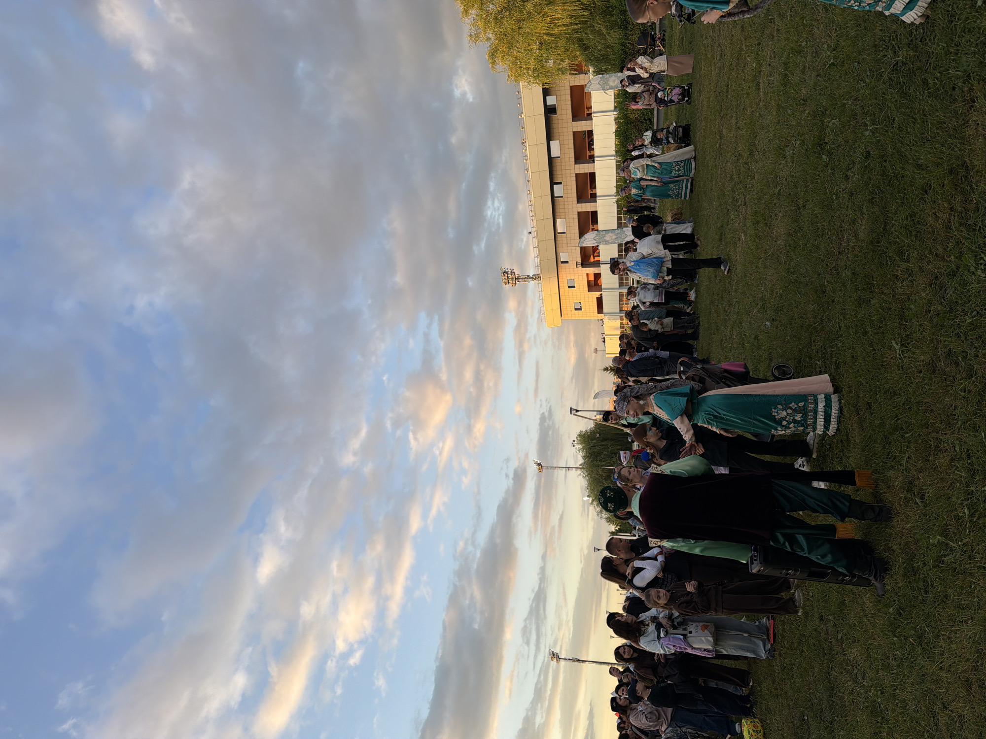In an outdoor grassy area, a diverse crowd of people of various ages are gathered under a cloudy sky with hints of sunset colors. Many individuals are dressed in traditional clothing, suggesting a cultural event or festival. Two women in ornate, teal and pink dresses stand out, engaged in conversation or performance. To the left, a man in a dark green vest and matching pants, adorned with gold tassels, stands near a black speaker. In the background, a modern building with a light-colored facade and large windows is visible, along with trees and lighting poles. The overall scene depicts a lively gathering during what appears to be twilight.