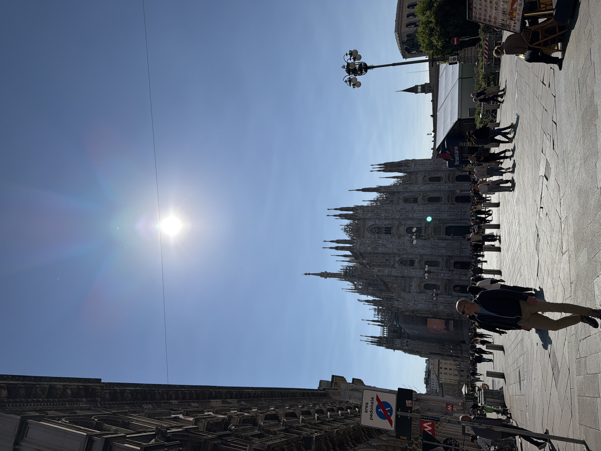 This media file captures a vibrant outdoor scene in Milan, Italy, centered around the majestic Milan Cathedral (Duomo di Milano). The image is taken on a bright, sunny day, indicated by the clear blue sky and the sun prominently featured in the upper left, creating a significant lens flare and long shadows that suggest late morning or early afternoon.
In the background, the intricate Gothic architecture of the Milan Cathedral dominates, with its numerous spires and detailed facade. To the left, a large building with classical, ornate stonework is visible. The foreground and midground feature a wide, paved public square, likely the Piazza del Duomo, bustling with people.
Numerous individuals are seen walking across the square. In the immediate foreground, a bald man wearing a dark jacket and khaki trousers walks towards the viewer. Further back, many other pedestrians are visible, some strolling casually, others forming small groups. On the right side of the square, an outdoor seating area with tables and chairs suggests a cafe or restaurant, with patrons seated and a menu board partially visible. A tall, multi-lamp street light stands near this area.
Notable objects include a blue circular sign with a red diagonal line through a pedestrian symbol, indicating a "pedestrian area" ("area pedonale" in Italian). Below it, a red 'M' symbol is visible, likely denoting a metro station entrance. A thin, dark line or wire stretches across the sky, passing through the sun's glare.
The overall atmosphere is lively and typical of a busy city square on a pleasant day, with people enjoying the outdoor environment.