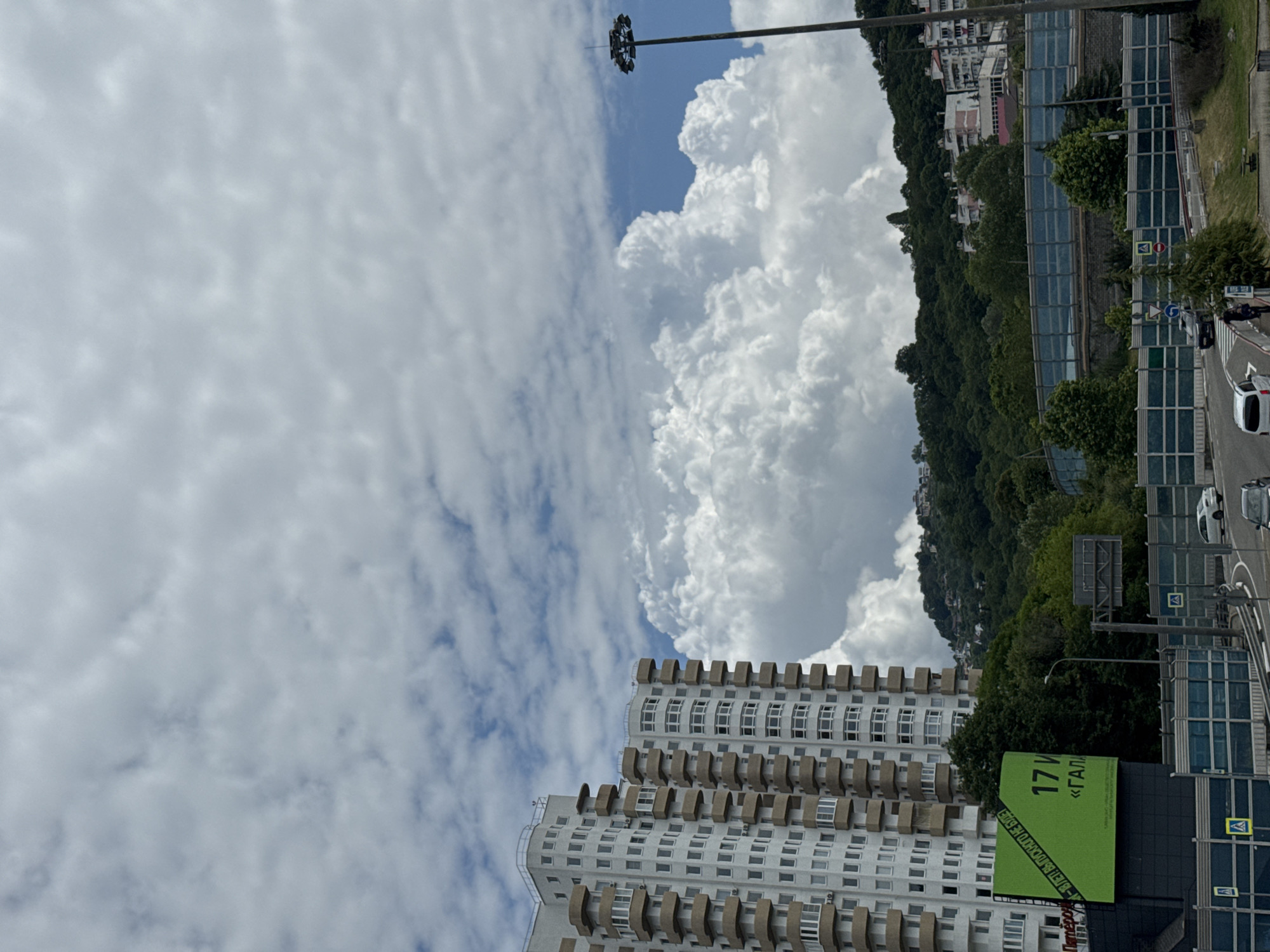 This image captures a panoramic view of a cityscape in Sochi, Russia, under a vast, cloudy sky. In the foreground, a multi-story apartment building with a distinct beige-trimmed facade and numerous balconies dominates the left side of the frame. Adjacent to the building is a large, bright green billboard displaying Russian text, including "17 И" and "«ГАЛЛ».
The middle ground features a lush, green hillside covered with dense trees, dotted with residential buildings and a tall, dark lamppost equipped with multiple lights on the right. A highway with protective sound barriers runs horizontally across the scene. Several vehicles are visible on the road, including cars and what appears to be a white van.
In the background, the sky is filled with dramatic, voluminous white clouds that suggest a dynamic weather system. The overall impression is of a coastal city nestled against a verdant, hilly landscape, bathed in diffused sunlight.