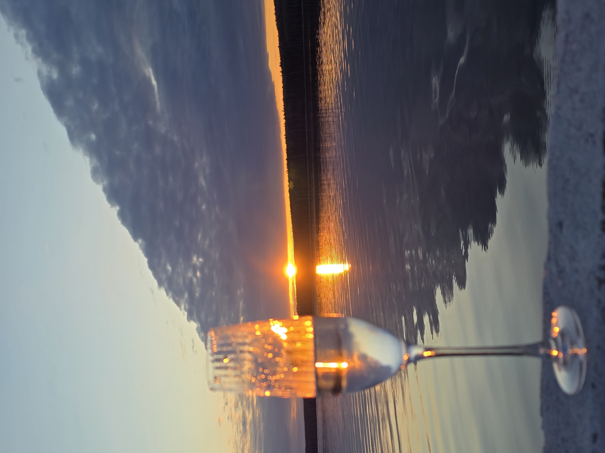 A horizontally oriented image depicts a sunset over a lake, with a champagne flute in the foreground. The lake extends into the distance, reflecting the orange and yellow colors of the setting sun. A dark line of trees is visible on the horizon. In the foreground, a champagne flute, positioned with its base on the right, is filled with a liquid that reflects the colors of the sunset. The image is rotated. This scene is likely located near the city of Chaikovsky in Russia, based on previous context. The cloud near the horizon suggests a storm coming.
