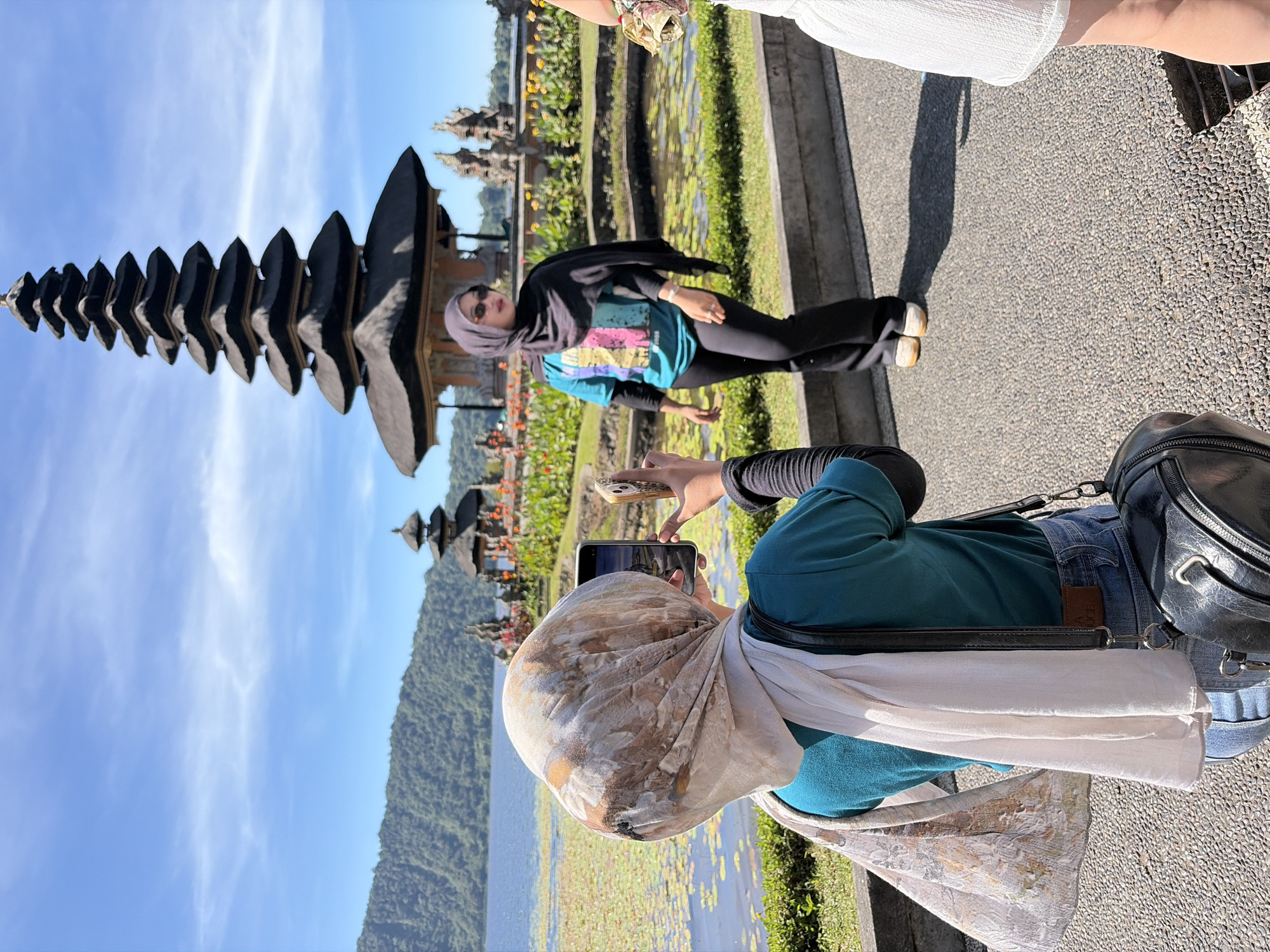 In Baturiti, Indonesia, on a sunny day, two women are photographed at the Pura Ulun Danu Bratan temple complex. The primary subject, in the foreground and from behind, is wearing a teal shirt, denim jeans, and a patterned headscarf with a white scarf draped over it. She is holding a smartphone in her right hand and another phone in her left hand, seemingly taking a picture of the second woman. A black leather bag is slung over her left shoulder. The second woman, positioned in the mid-ground, faces the camera, wearing a black hijab, sunglasses, a blue and pink patterned t-shirt, and black pants. She is standing on a paved walkway near a body of water with lily pads. In the background, the iconic multi-tiered pagoda of the temple rises against a bright blue sky with scattered clouds. Hills covered in lush greenery are visible across the lake. Another, smaller temple structure is also visible in the background. A third person, only partially visible, stands on the far right, wearing a white skirt. The environment is a tourist attraction with paved paths, manicured grass, and a serene lake.