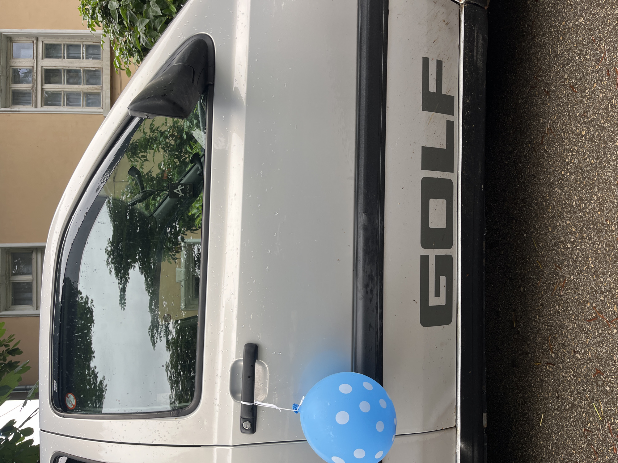 An outdoor, eye-level shot captures the side of a light-colored car, identified by the prominent "GOLF" text in dark gray on its lower panel. A blue balloon with white polka dots is tied with a white string to the car's black door handle. The car's surface, particularly the door and window, is speckled with water droplets, indicating recent rain or heavy dew.
The car's window reflects tall green trees and parts of a building. In the background, a light-colored building with an old-fashioned, multi-paned window is visible. The ground next to the car appears to be dark asphalt or pavement. The scene is illuminated by daylight. No people are present, and the main activity suggested is the presence of the balloon, perhaps for a celebration.