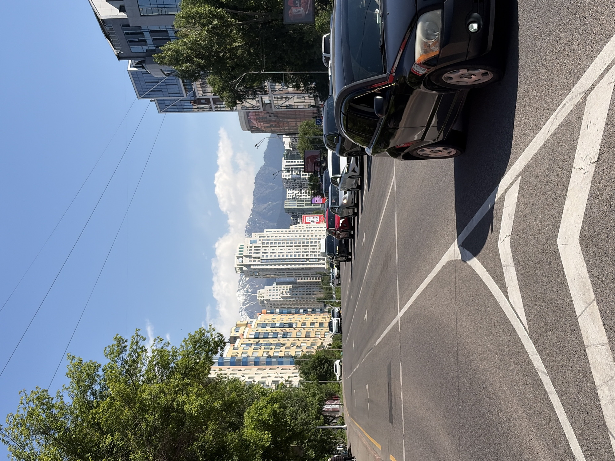 A street in Almaty, Kazakhstan, with buildings and mountains in the background. Cars are parked along the right side of the road. The road has white lines painted on it. Trees are visible on the left side of the road. The sky is blue with some clouds.
