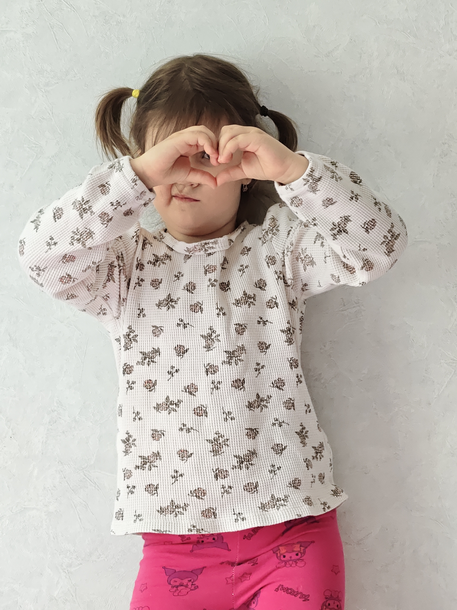 A young girl, likely a toddler or preschooler, stands centered against a light-colored, subtly textured wall indoors in Chaikovsky, Russia. She has brown hair styled in two pigtails, each secured with a yellow elastic. The girl is wearing a long-sleeved, off-white or cream-colored shirt with a waffle-knit texture. The shirt is patterned with small, stylized floral or leaf motifs in shades of brown and dark green, and features a subtle ruffled collar. She is also wearing bright pink leggings adorned with repeating cartoon characters, specifically Kuromi, and small star patterns. The word "Kuromi" is visible on the leggings.
The primary activity depicted is the girl making a heart shape with both hands held up in front of her face, peering playfully through the opening of the heart gesture. Her facial expression, partially obscured by her hands, appears gentle. The lighting suggests an indoor setting, likely during the daytime. The overall mood is one of affection and innocence.