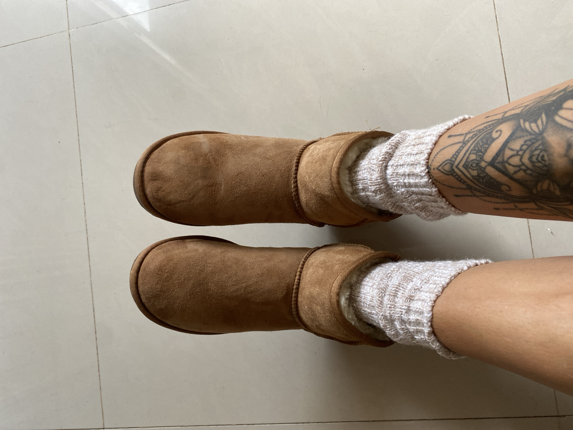 An overhead, full-shot image displays the lower legs and feet of a person, likely indoors on a light-tiled floor in Mũi Né, Vietnam. The subject is wearing a pair of light brown, mid-calf UGG-style boots, made of a suede-like material with visible, fluffy light-colored lining at the top. Inside the boots, light beige or off-white knitted socks with a textured pattern are worn, pulled up above the ankle.
The left leg is adorned with a large, intricate black ink tattoo, possibly a mandala or geometric floral design, covering a significant portion of the calf/shin area. The right leg is bare, showing smooth skin. The floor consists of large, light-colored, possibly ceramic or porcelain tiles with faint grout lines. Bright, likely natural, overhead lighting illuminates the scene, creating reflections on the shiny floor surface in the upper left, suggesting it is daytime. The boots appear somewhat worn, indicating they are well-used. No specific activity is depicted other than the person standing or sitting with their feet on the floor. There is no visible text in the image.