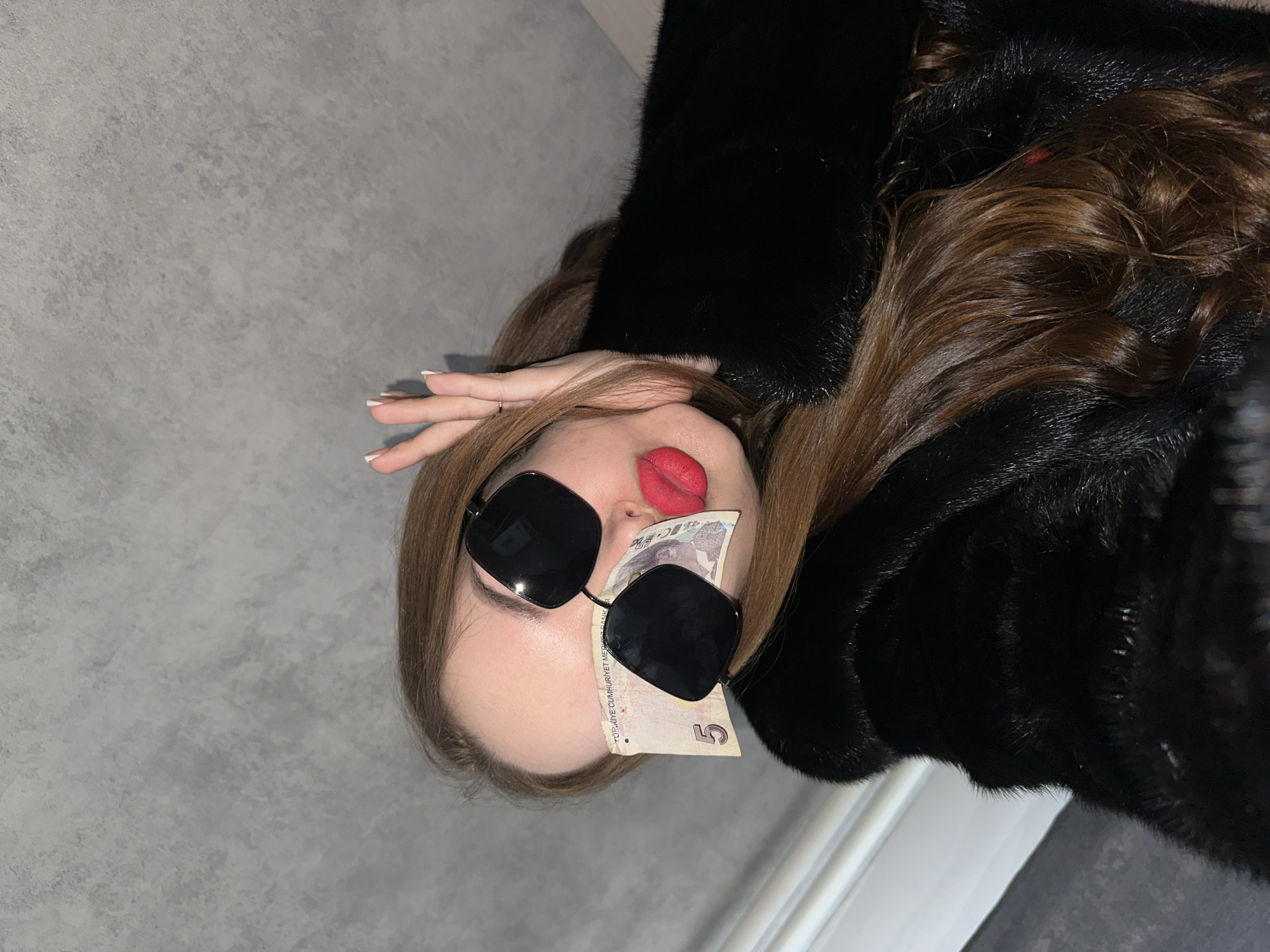 A woman is lying on a grey floor, wearing a black fur coat and dark sunglasses. A five Turkish lira bill is partially tucked under the sunglasses and covering her nose. Her lips are painted red. She is positioned at an angle, with her head tilted towards the top left of the frame. Her right hand is raised near her forehead. The location is in the city of Chaikovsky, Russia.