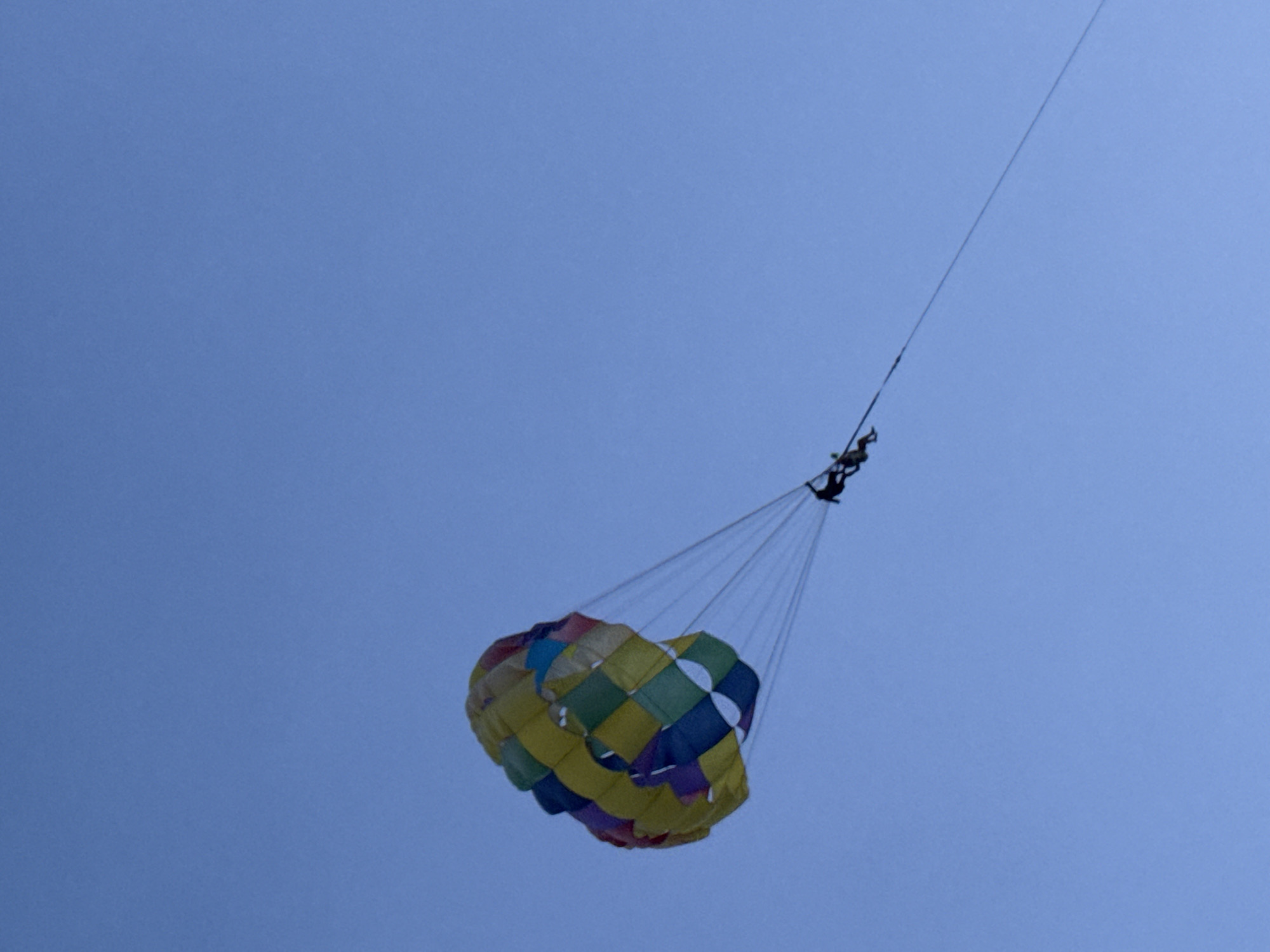 A colorful parasail, patterned with yellow, blue, green, and purple squares, is suspended against a clear blue sky. Two figures are visible, harnessed to the parasail, with one appearing to be seated and the other further back and slightly lower. The parasail is being towed by an unseen force, indicated by the taut rope extending diagonally upwards and to the right, disappearing out of frame. The scene suggests a recreational activity taking place in an open, outdoor environment. This activity is commonly observed at beaches in Karon, Thailand, known for water sports and aerial adventures.