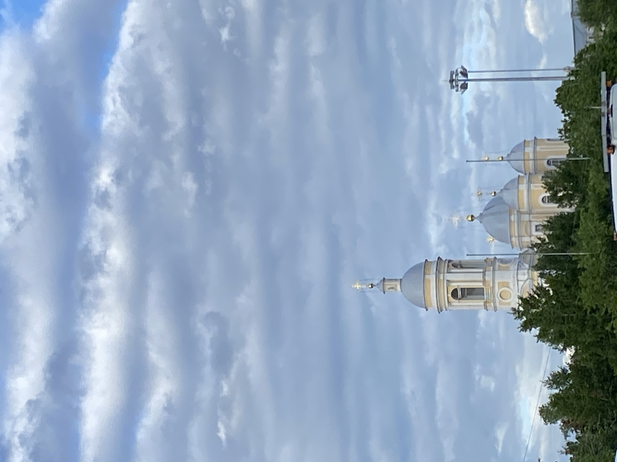 The image shows a view of the Smolny Cathedral in Saint Petersburg, Russia. The architecture of the cathedral, with its characteristic white and gold domes, is prominently visible against a sky filled with clouds. The scene also includes trees and a light pole, giving context to the cathedral's surroundings.