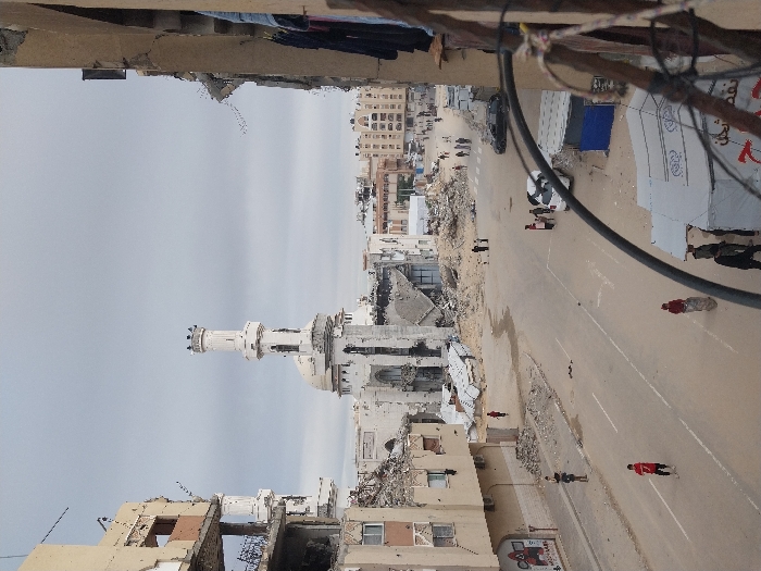 This image depicts a street scene in Khan Yunis, Palestinian Territory, under an overcast sky. The perspective is from an elevated vantage point, possibly from a damaged building on the right.
In the foreground, the lower right is partially obscured by dark cables or wires. Below these cables, two white tents and a smaller blue tarp structure are visible on the roadside, with approximately five individuals standing near them. One person in a red shirt is partially visible near a sack.
The midground shows a paved street with white lane markings, extensively covered with debris, rubble, and dirt, along with several wet patches or puddles. Multi-story buildings lining both sides of the street exhibit severe structural damage, including exposed rebar, collapsed sections, and shattered windows. A mural or graffiti is visible on a lower wall of the damaged building on the left. Approximately eight individuals or small groups are present on the street and sidewalks: one person in a red shirt walks on the left, another in a red shirt stands near a sack, a group of four people gathers near a white car on the right, and other pedestrians are walking further down the street. A black car is parked further down the street on the right.
The background is dominated by a large mosque featuring two prominent white minarets and a dome. While the minarets and dome show visible signs of wear and minor damage, the main mosque structure behind them appears heavily damaged or partially collapsed, with a substantial pile of rubble at its base. Beyond the mosque, additional urban buildings, some appearing less damaged, extend into the distance.