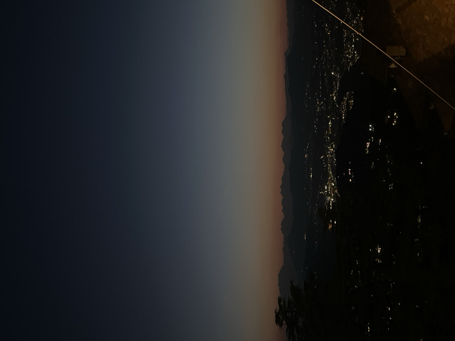 The image depicts a panoramic twilight or night view captured from an elevated position in Brunate, Italy. The upper two-thirds of the frame show a gradient sky, transitioning from dark blue-black at the top to a subtle orange-red hue near the horizon. A continuous, dark, jagged silhouette of a mountain range extends across the mid-ground, separating the sky from the urban landscape below. Beneath the mountains, a dense concentration of artificial lights illuminates a sprawling city, identified as Como, with individual streetlights and building illuminations visible as bright points against the dark terrain. The immediate foreground on the left is occupied by the dark silhouette of deciduous foliage or a tree. On the lower right, a dark, angled structure, consistent with a building roofline or wall, is partially visible, from which a light-colored, diagonal line, possibly a wire or support cable, extends upwards towards the center of the frame. No individuals, explicit actions, or interactions are visible within the scene.
