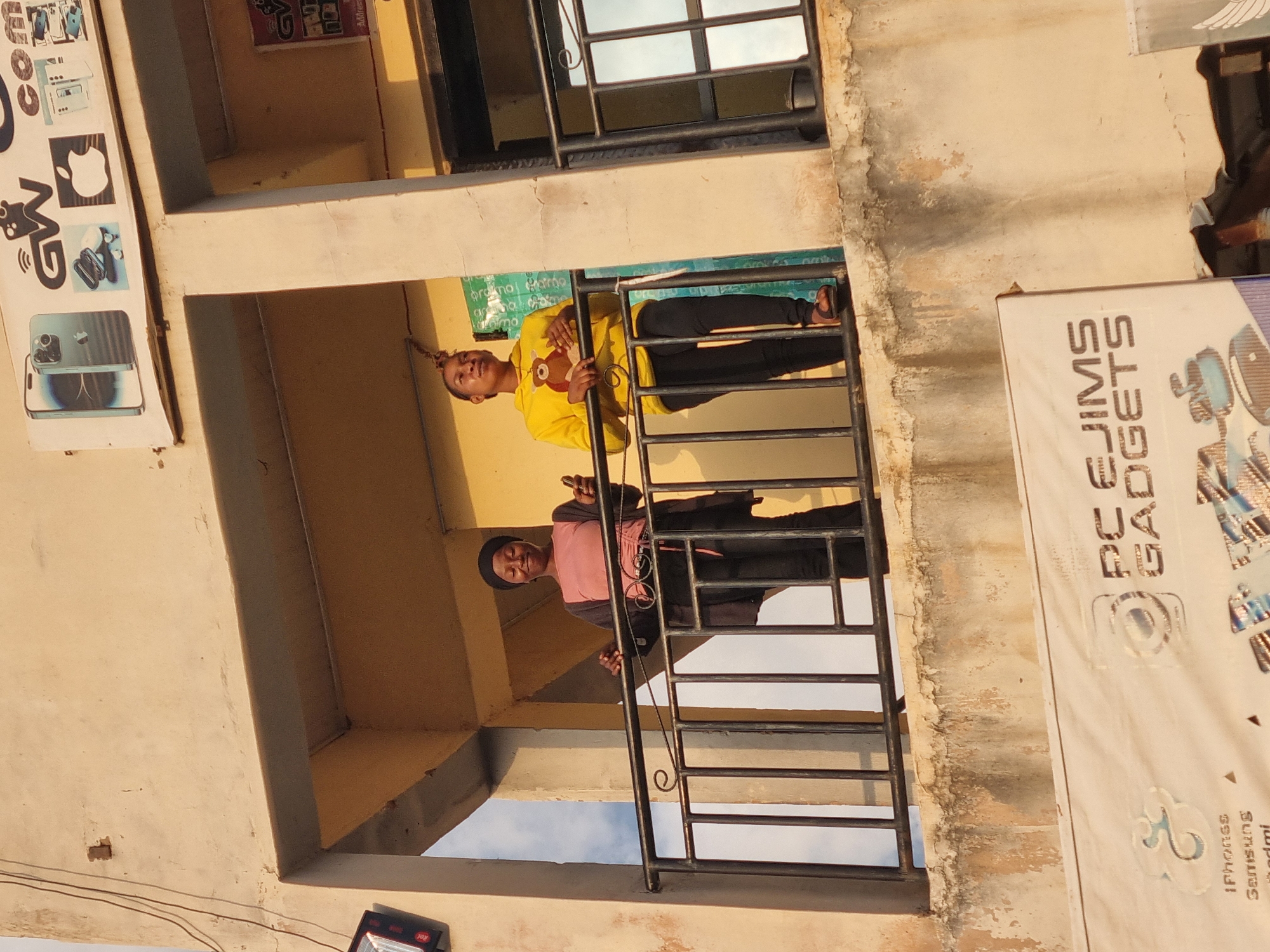 The image depicts a building in Jos, Nigeria, with two women standing on a balcony. One woman is wearing a yellow top and black pants, while the other is wearing a pink top and black pants. A sign for "PC FJIMS GADGETS" is visible on the right side of the image. Another sign is visible on the left side. The building appears to be multi-storied. The sky is visible through the balcony railing.