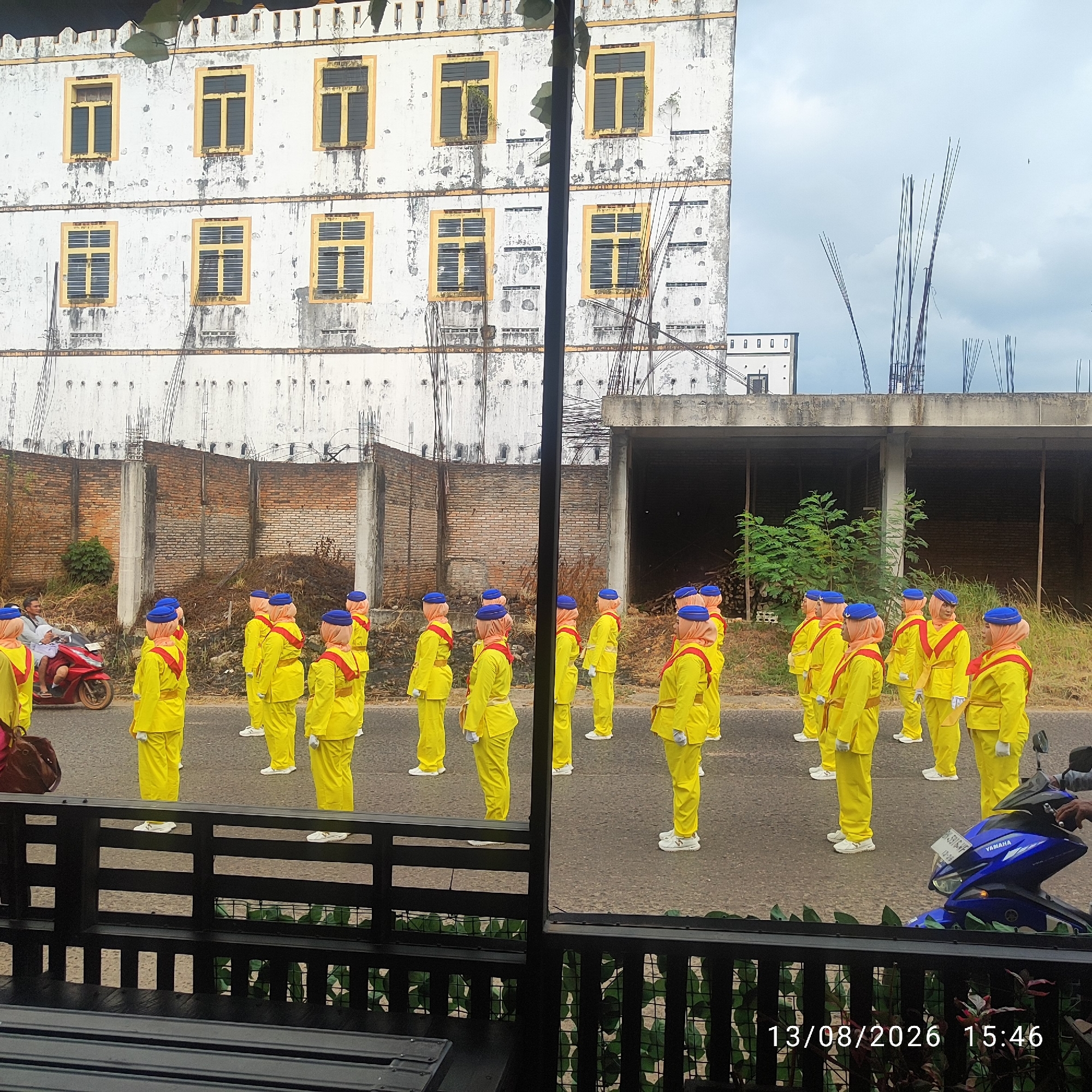 In Buding, Indonesia, a group of individuals in bright yellow uniforms with blue caps and red sashes stand in formation on a paved road. Some are facing forward, while others are looking to the side. A man is seated on a red motorcycle to the left, looking towards the group. In the background, a large, weathered building with multiple windows and peeling paint dominates the scene. Beyond the building, construction scaffolding and brick walls are visible, suggesting an urban or developing area. The sky is overcast with some visible clouds. A black metal fence partially obscures the foreground, with a portion of what appears to be furniture visible behind it. The timestamp "13/08/2026 15:46" is overlaid on the bottom right of the image.
