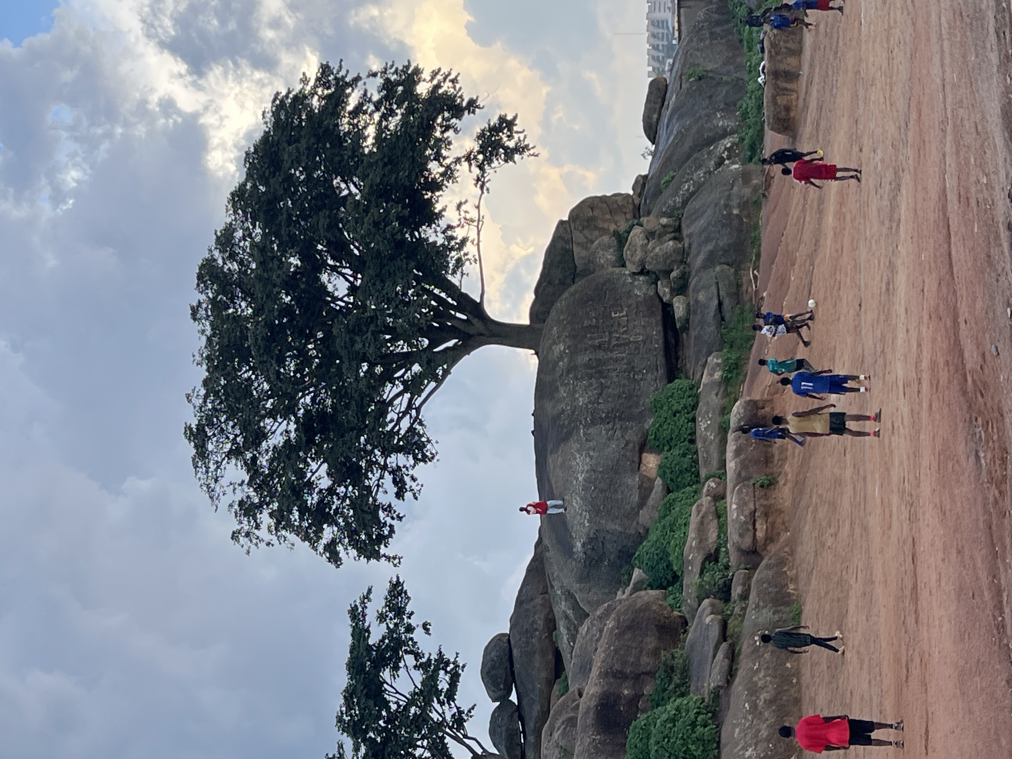 The image depicts a group of young men playing soccer on a dusty, reddish-brown field at the base of a large rocky outcrop in Jos, Nigeria. A prominent, mature tree with dark green foliage stands atop the highest point of the outcrop, casting a shadow over the rocks. Several large boulders are scattered across the landscape, some covered with green vegetation. In the background, a few buildings are visible, suggesting an urban or semi-urban setting. The sky is overcast with scattered clouds, with sunlight breaking through in the upper right quadrant. The players are dressed in casual athletic wear, with some wearing distinctive jerseys. One individual, wearing a red top and blue shorts, is captured mid-action, appearing to be running with the ball. Other individuals are positioned around the field, some actively engaged in the game, while others observe. A person in a red shirt and white pants is standing on a rock formation, looking down at the field. The overall scene captures a moment of recreation and daily life in Jos.