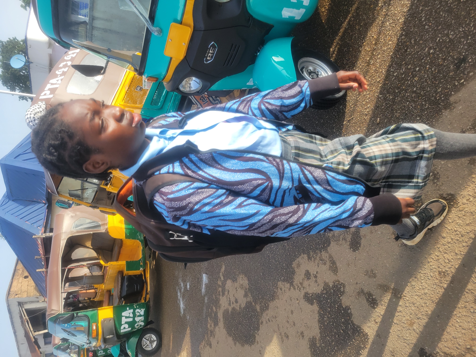 A child stands on a pavement next to a road, with "keke napep" auto rickshaws parked nearby. The child is wearing a blue and black patterned jacket over a lighter blue shirt, a plaid skirt, gray leggings, and black and gray sneakers. They have a backpack on. The surrounding environment includes other "keke napep" vehicles, some with the writing "PTA-433-Q12", indicating they are likely in Jos, Nigeria. The pavement appears to be wet in places.