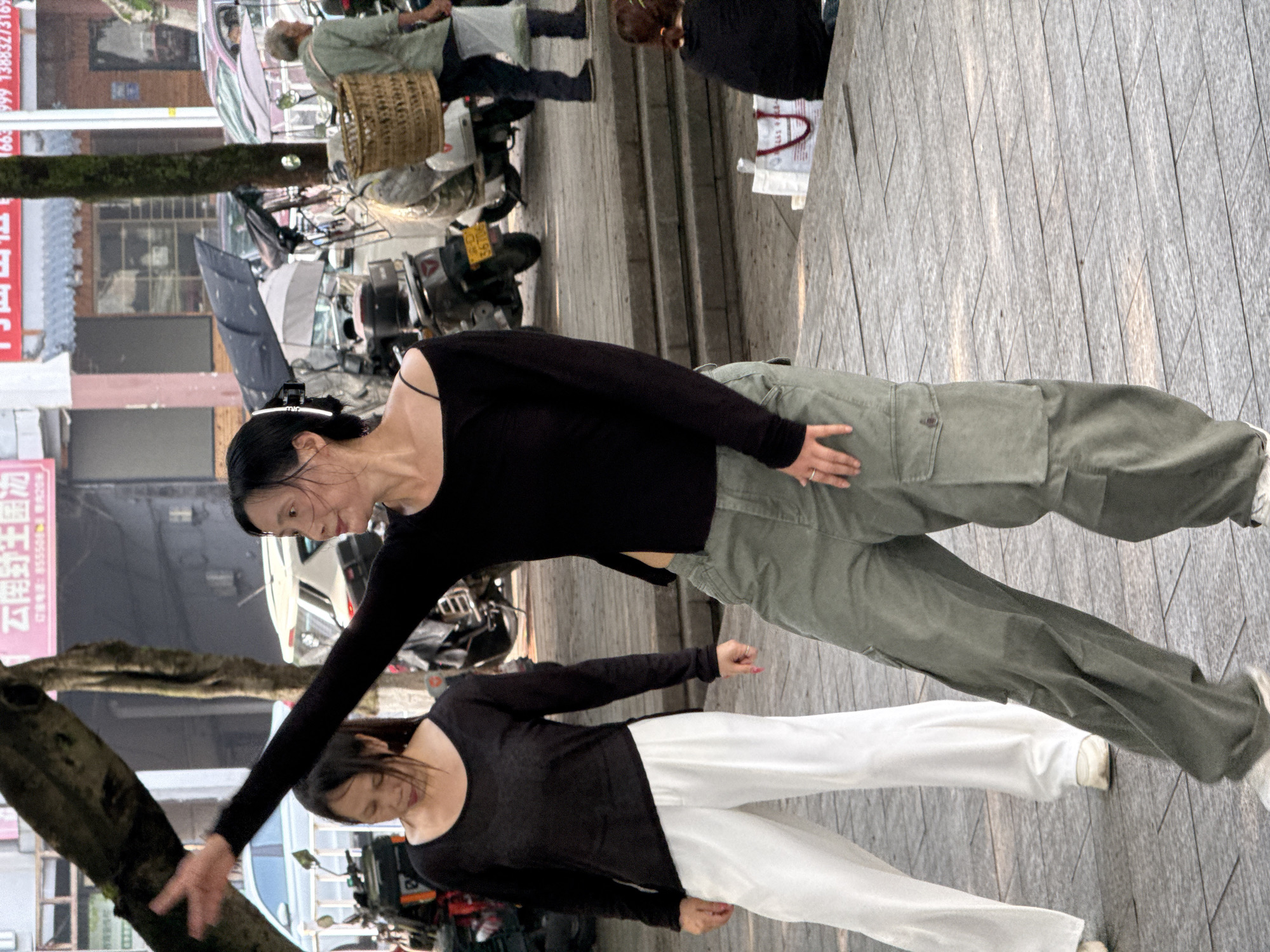 In Jiangjin, China, on a paved plaza with visible steps, two women are engaged in what appears to be a dance or exercise. The woman in the foreground, wearing a black off-the-shoulder top and olive-green cargo pants, is captured mid-movement with her right arm extended and hand open. Her left hand is placed on her hip. To her left and slightly behind, another woman in a black long-sleeved top and white wide-leg pants mirrors a similar stance, with her head bowed. The background reveals a street scene with parked motorcycles, a car, and buildings with signage. The lighting suggests it is daytime, possibly late afternoon, with sunlight casting shadows. The environment appears to be an urban public space.