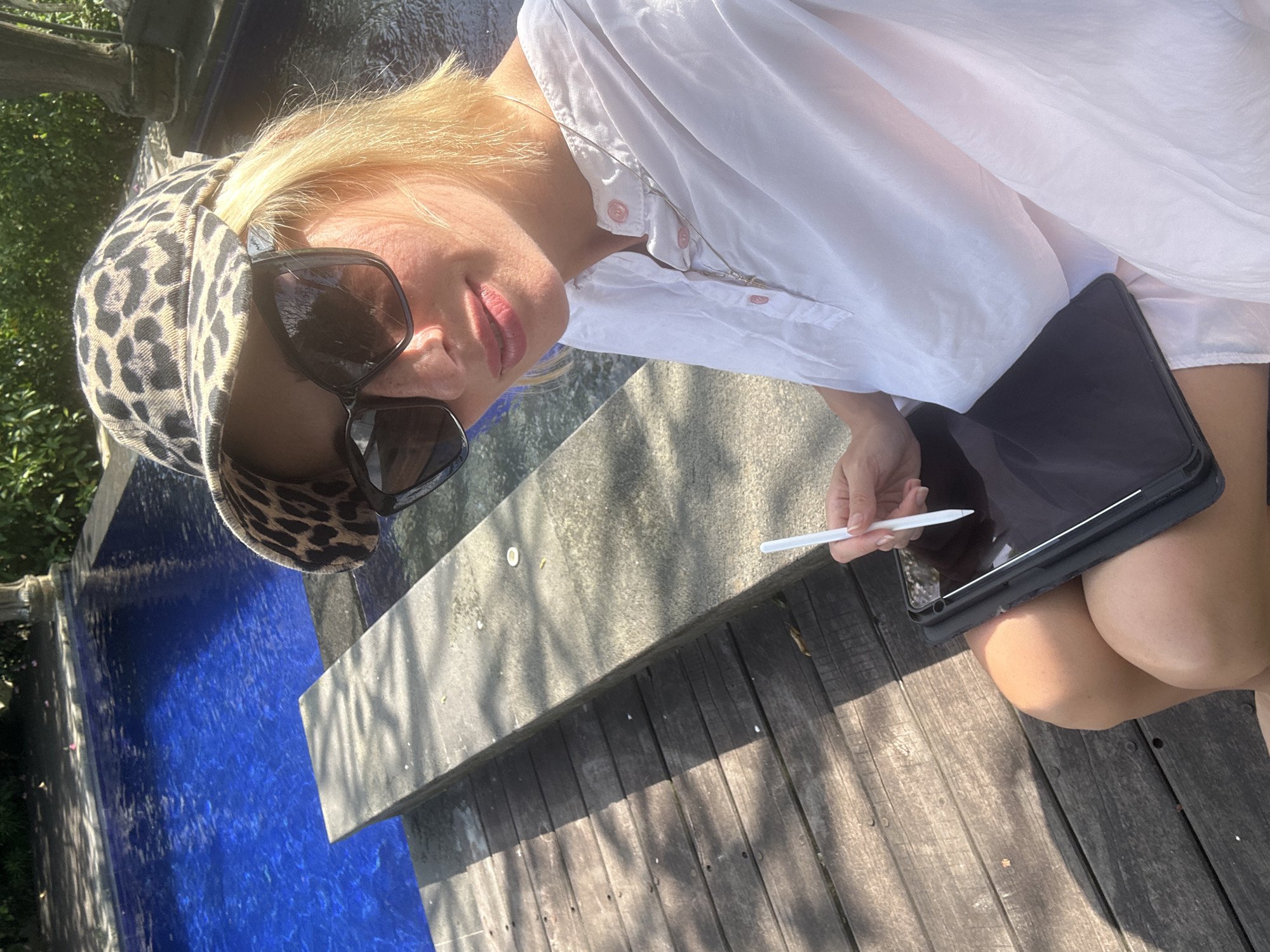 A blonde woman with sunglasses and a leopard print cap is sitting outdoors in Kediri, Indonesia. She is wearing a white button-down shirt, holding a stylus and a tablet. The backdrop includes a blue swimming pool, stone, and wooden decking. The scene is lit with bright sunlight.