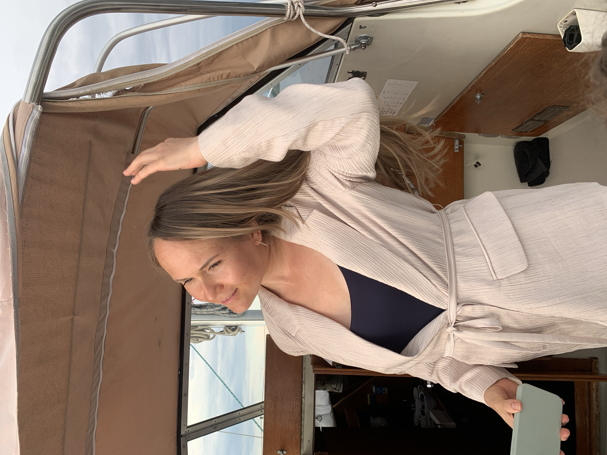 In Chaikovsky, Russia, a woman with blonde hair, wearing a light-colored, belted jacket over a dark top, stands on a boat. She is looking upwards and to the left, with her right hand touching her hair. The boat's interior is visible, with wooden paneling and a canvas covering overhead. The sky is cloudy.