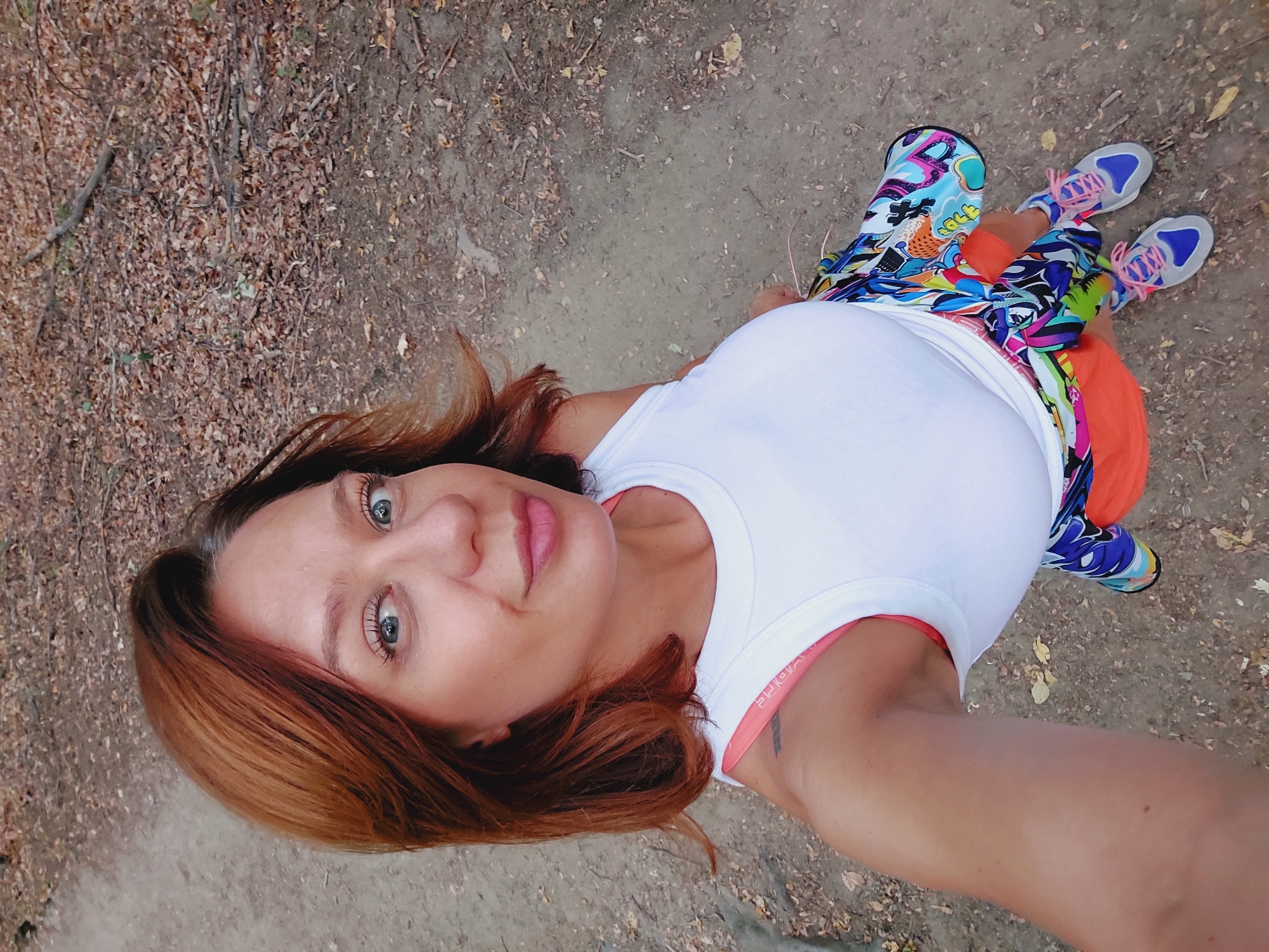 A woman with reddish-brown hair is standing on a dirt path covered with fallen leaves, possibly in the city of Bistritsa, Bulgaria. She is wearing a white tank top, brightly colored leggings with a graphic pattern, and athletic shoes with pink laces. An orange jacket is tied around her waist. The background consists of trees and foliage, suggesting an outdoor, natural setting. She is looking up and slightly to the right, taking a selfie.
