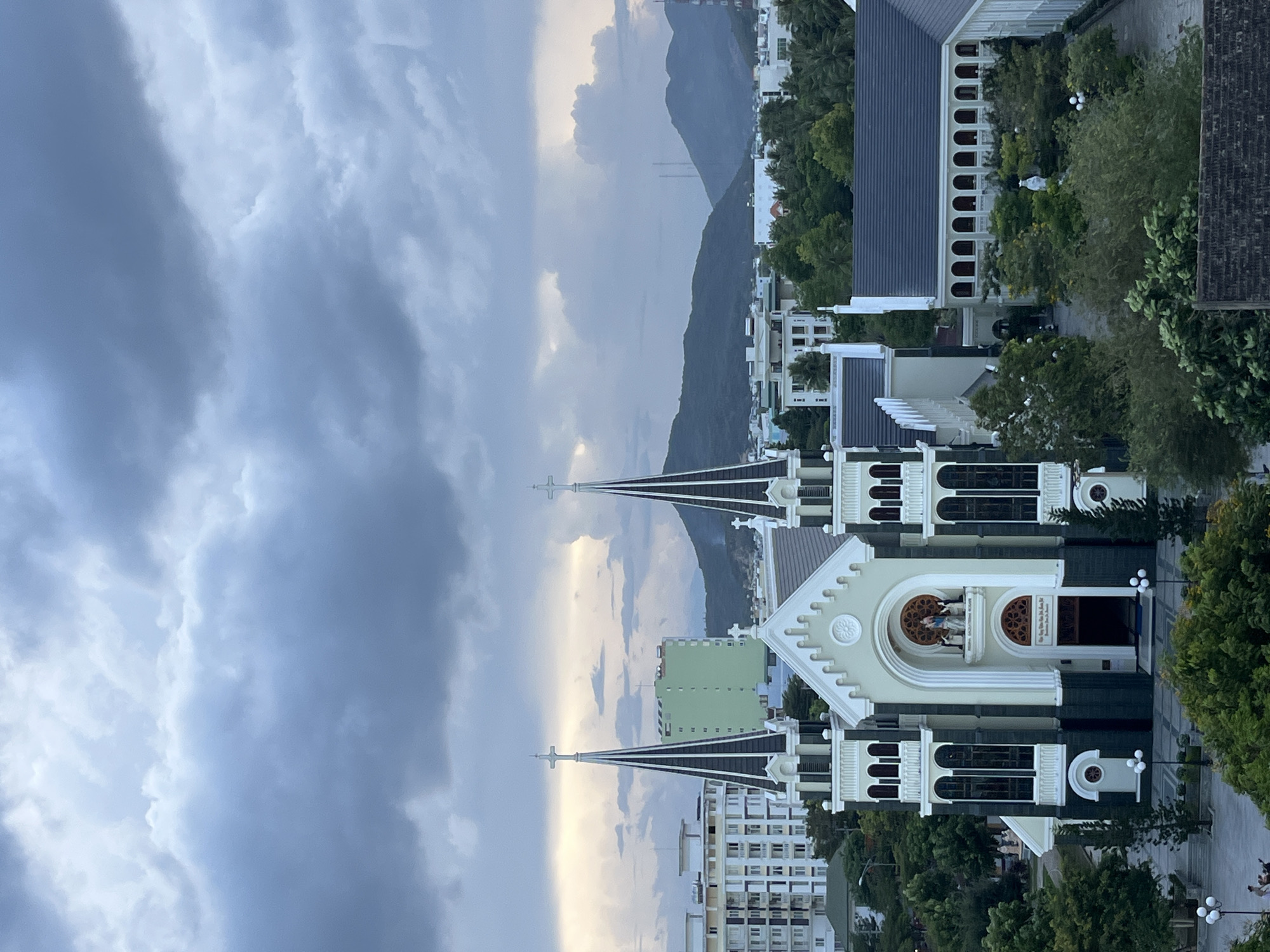The image captures an elevated view of Nha Trang Cathedral, also known as the Stone Church, in Bac Nha Trang Ward, Vietnam. The church, characterized by its white and dark gray neo-Gothic architecture, features spires with crosses on top. The facade includes a sculpture of a religious scene. The surrounding area comprises trees, buildings, and a street with streetlights. In the background, a mountain range is visible under a cloudy sky.