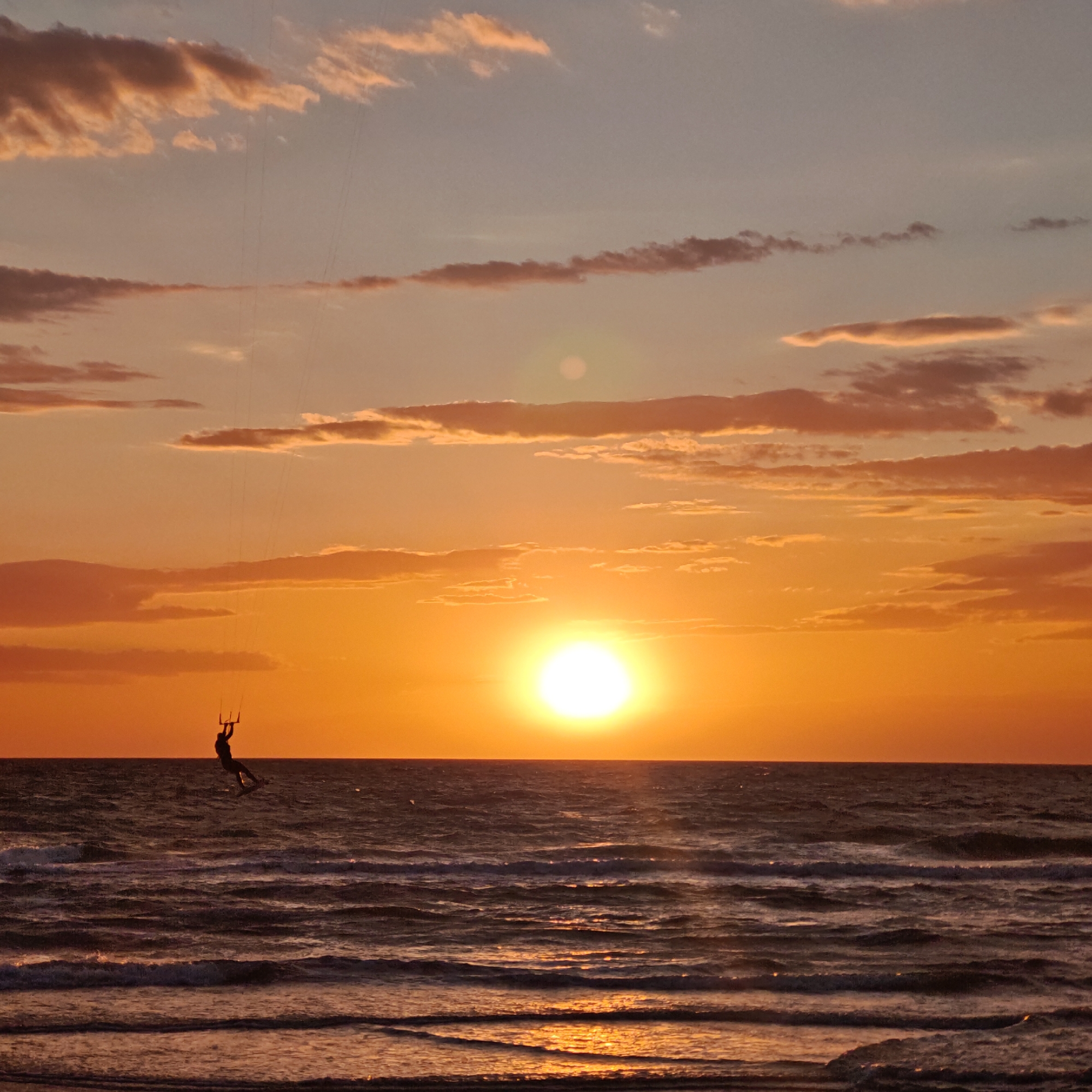 A wide shot captures a kitesurfer in mid-air against the backdrop of a sunset over the water. The location is городское поселение Щелкино, Ukraine. The sun sits just above the horizon, casting a bright glow that reflects on the water. The sky is filled with clouds tinged with orange and pink hues. The kitesurfer is silhouetted against the bright sun, with the kite lines extending upwards. The water is choppy with small waves.