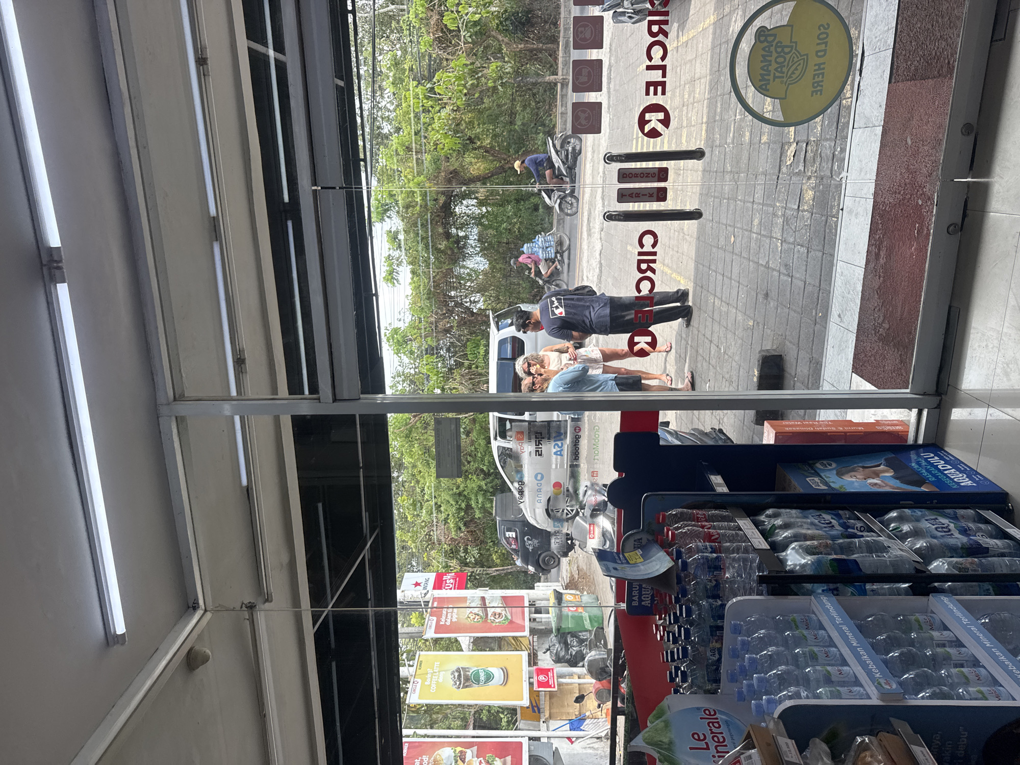 An exterior shot through the glass doors of a Circle K convenience store in Kuta Selatan, Indonesia, shows an outdoor scene with several people and vehicles. Inside the store, shelves stocked with bottled water are visible. The glass doors have red "CIRCLE K" logos and some text in Indonesian. Outside, three people are standing near the entrance, two women and one man. A white van is parked on the side of the road, and two men on motorcycles are visible further down the street, one carrying several large water jugs. Trees and dense foliage line the background. A yellow circular sign with a banana and the text "BANANA BOAT" is visible on the glass door to the right. The ground outside is paved with square tiles.