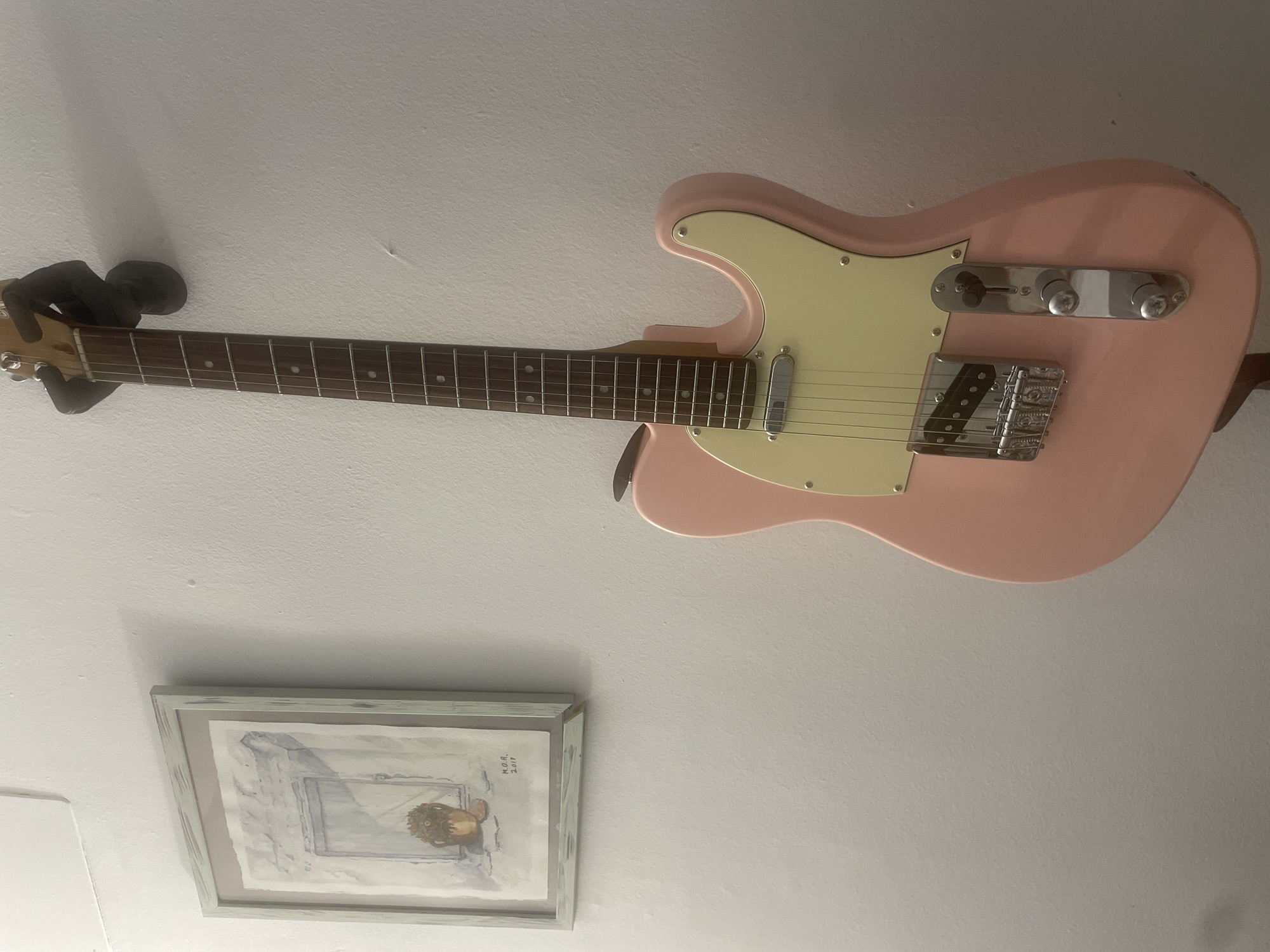 A pink electric guitar is mounted on a wall in a room. To the left of the guitar, a framed watercolor painting of a window with a potted plant is also mounted on the wall. The guitar appears to be a Telecaster style guitar with a light yellow pickguard and chrome hardware. The wall is painted white and shows some texture. The lighting is dim, creating shadows on the wall. The image does not contain any information about the location within the city of Atarfe, Spain.