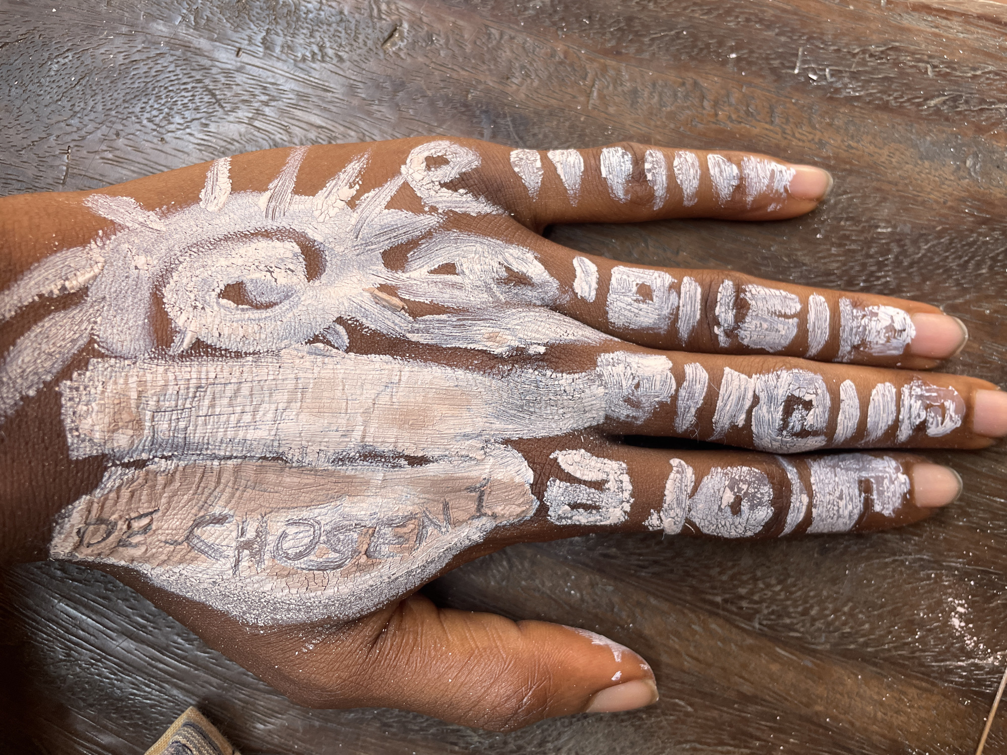 The image displays a human hand, likely belonging to a person of color, resting on a textured dark brown wooden surface. The hand is adorned with white paint, applied in various patterns and words.
On the back of the hand, centered, is a distinctive sun-like or floral design. Below this design, faint text reading "BE CHOSEN" is visible, appearing to be written in pencil or scratched onto the skin or dried paint, with some thicker white paint marks over it.
Each finger features vertically written words in white paint: "LIVE" on the index finger, "LOVE" on the middle finger, "HOPE" on the ring finger, and "JOY" on the pinky finger. The paint shows signs of drying and cracking in places, suggesting it is a temporary body paint. The fingernails are natural.
The setting appears to be indoors or in a shaded outdoor area, with even lighting. The wooden surface beneath the hand has a prominent grain pattern. This body art, potentially a form of cultural expression or personal affirmation, given the positive words, is depicted in Onitsha, Nigeria.