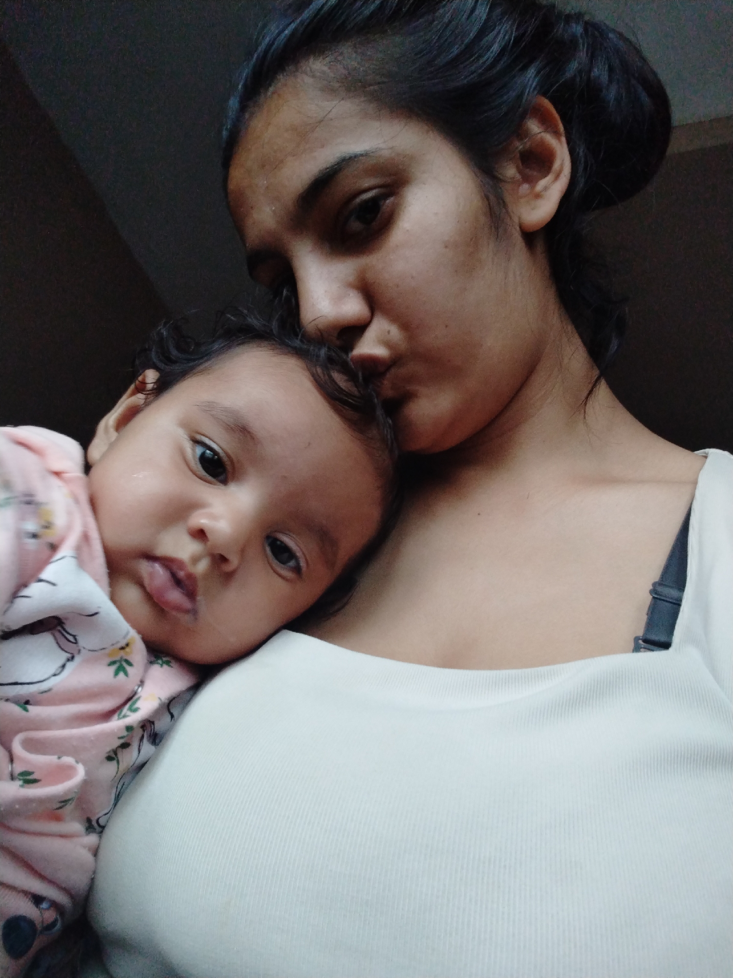 A medium shot depicts a dark-haired woman holding a baby. The woman is positioned behind the baby, and she is kissing the baby's forehead. The baby is wearing a pink onesie with a light pattern. The woman has her hair up in a bun and is wearing a white top. The background is dark and indistinct. The location is Unknown, Unknown.