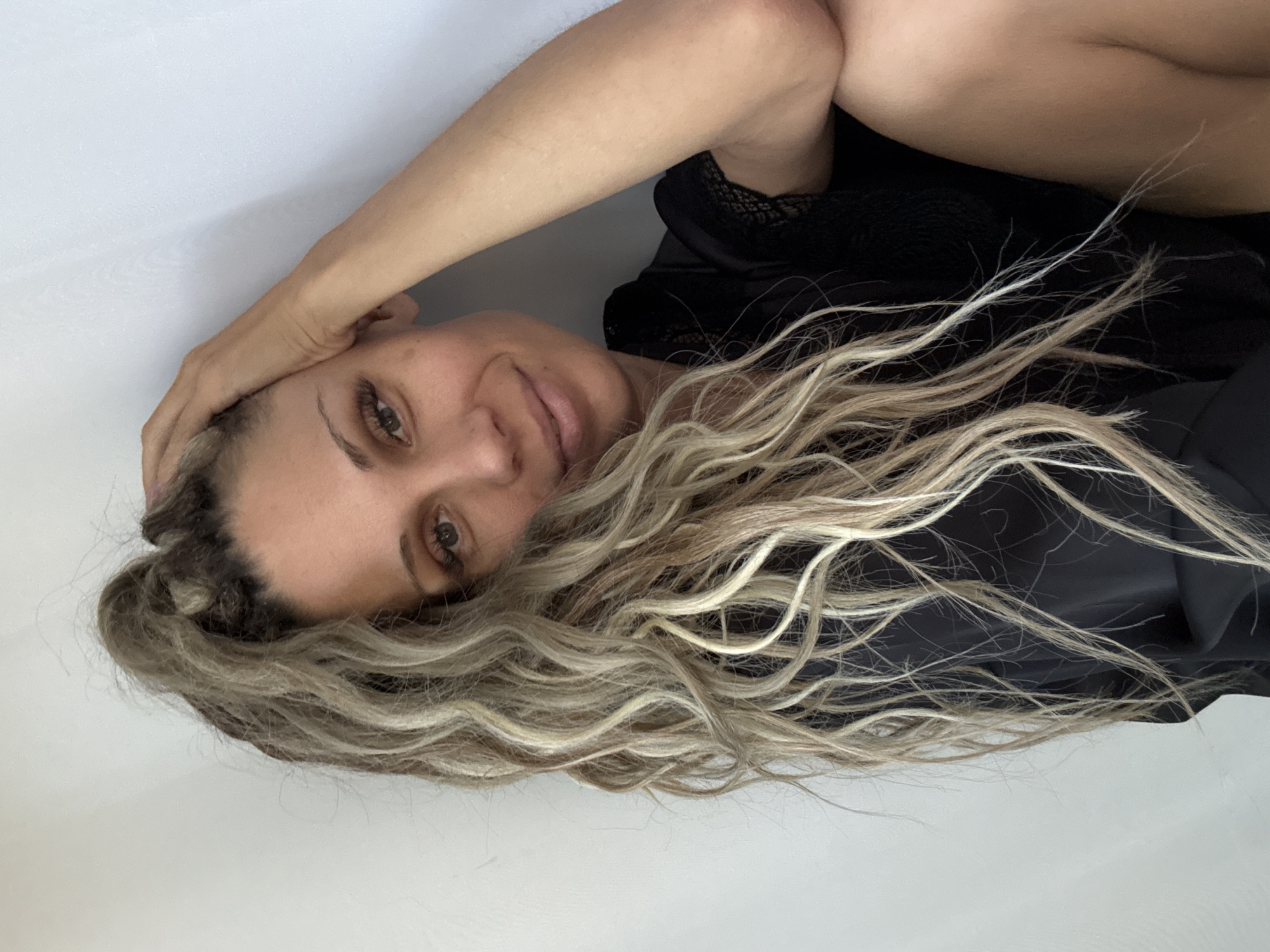 A woman with long, wavy blonde and brown hair pulled into a high ponytail is positioned in the lower right of the frame, leaning against a plain white wall. She is wearing a black robe with lace trim. Her left hand is raised and resting on her head. She is looking towards the camera with a slight smile. The background is a solid, light-colored wall. There is no indication of the city of Rusanovo, Russia in this image.