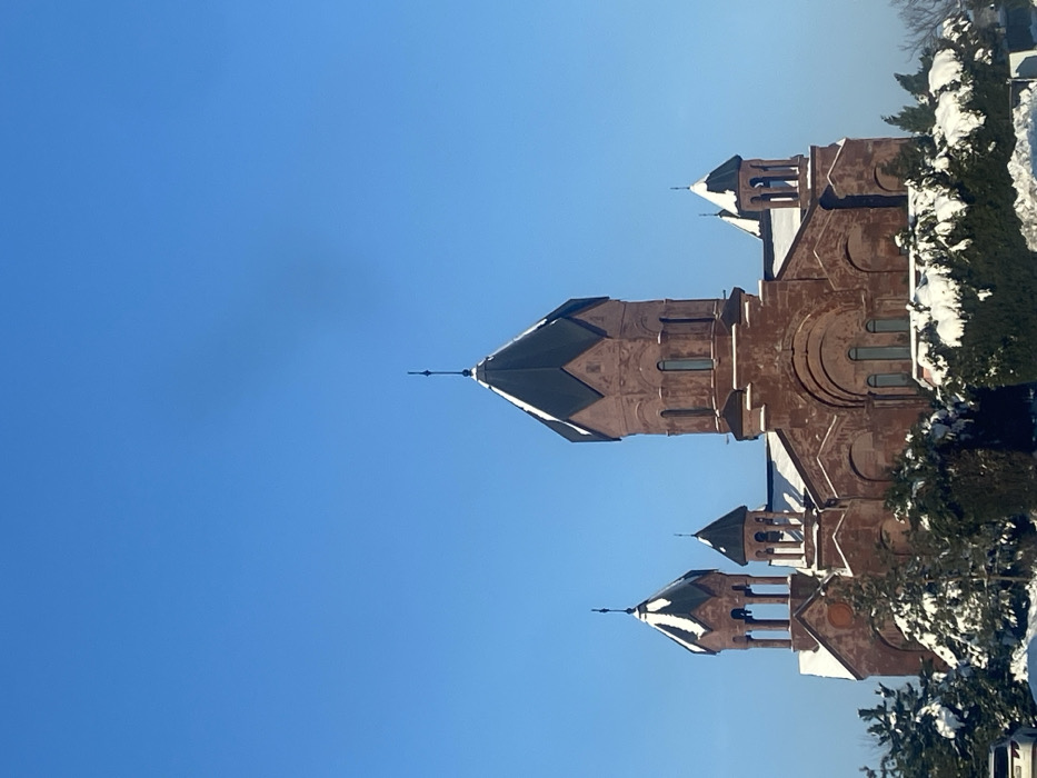 The image displays an exterior view of the Surb Astvatsatsin (Holy Mother of God) Armenian Church in Gay-Kodzor, Russia, under clear winter conditions. The church is constructed of reddish-brown stone or brick masonry, featuring a prominent central octagonal drum capped by a dark, conical roof with a finial. Three additional, smaller domes with dark conical roofs and finials are visible on the structure, partially covered with a thin layer of snow. Arched windows are present on the church's facades and drums. The ground and surrounding evergreen bushes and trees are heavily blanketed in white snow. The sky is a clear, cloudless blue. A small portion of a light-colored vehicle is visible in the bottom-left corner, obscured by snow-covered foliage. No individuals are depicted.