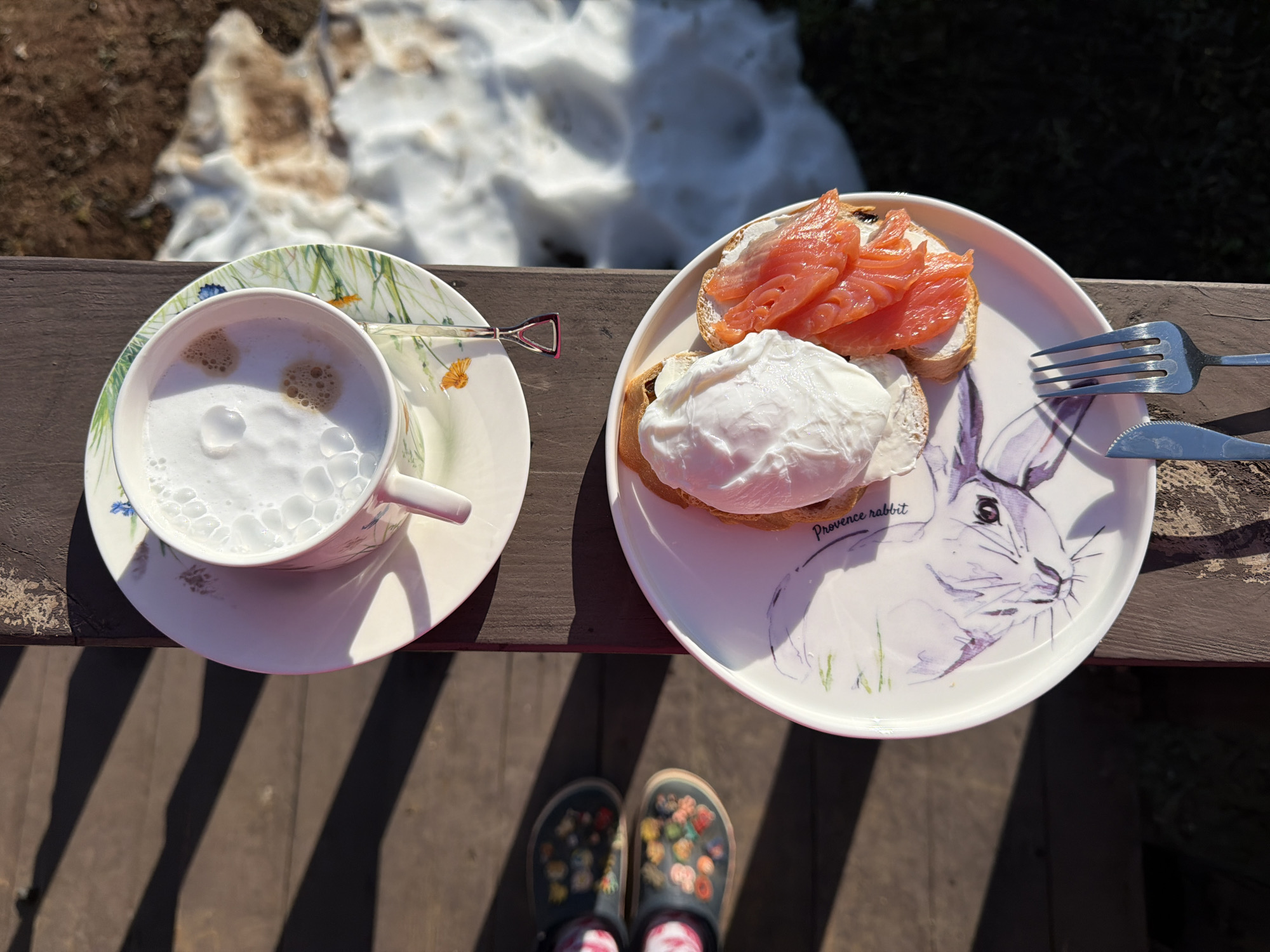 The image presents an overhead view of an outdoor breakfast or brunch setup in Chaikovsky, Russia, seen from the perspective of the person about to enjoy it. The person's feet, clad in black clogs with colorful floral embellishments and patterned socks, are visible at the bottom of the frame.
On the left, a white teacup with a delicate floral pattern of green stems and small orange and blue flowers sits on a matching saucer. It contains a foamy, white beverage, likely a latte or cappuccino, with a decorative spoon resting on the saucer.
To the right, a round white plate holds two pieces of toast. One is topped with several slices of pink salmon and a white spread, while the other features a perfectly poached egg. The plate itself is adorned with a purple-toned illustration of a rabbit and the text "Provence rabbit". A fork and a serrated knife are placed beside the plate.
The meal is arranged on a dark wooden surface, possibly a deck or outdoor table. The background shows patches of melting snow mixed with bare ground, suggesting an early spring or late winter setting. Strong, elongated shadows cast by the bright sunlight across the wooden surface indicate a clear morning or early afternoon. The scene evokes a sense of calm and enjoyment of a meal outdoors on a cool, sunny day.