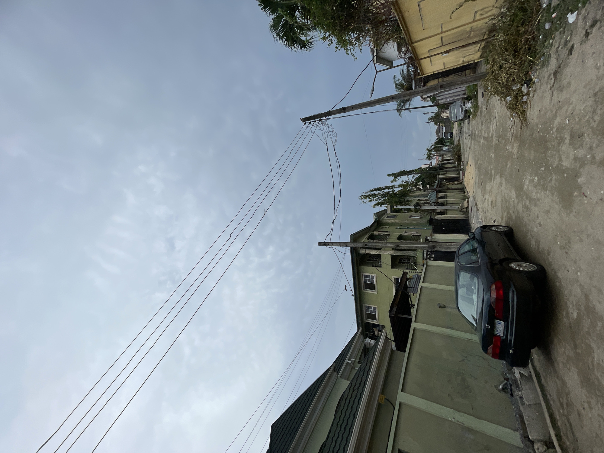 The image depicts a street scene in an urban area. A dark-colored sedan is parked on the right side of the street, close to a light green painted wall. The street itself appears to be concrete and shows signs of wear. Buildings of various heights and architectural styles line the street, some painted in light green. Overhead, power lines stretch across the sky, connecting to utility poles. The sky is overcast with grey clouds. The exact location within the city of Unknown, Unknown is not discernible from the image.