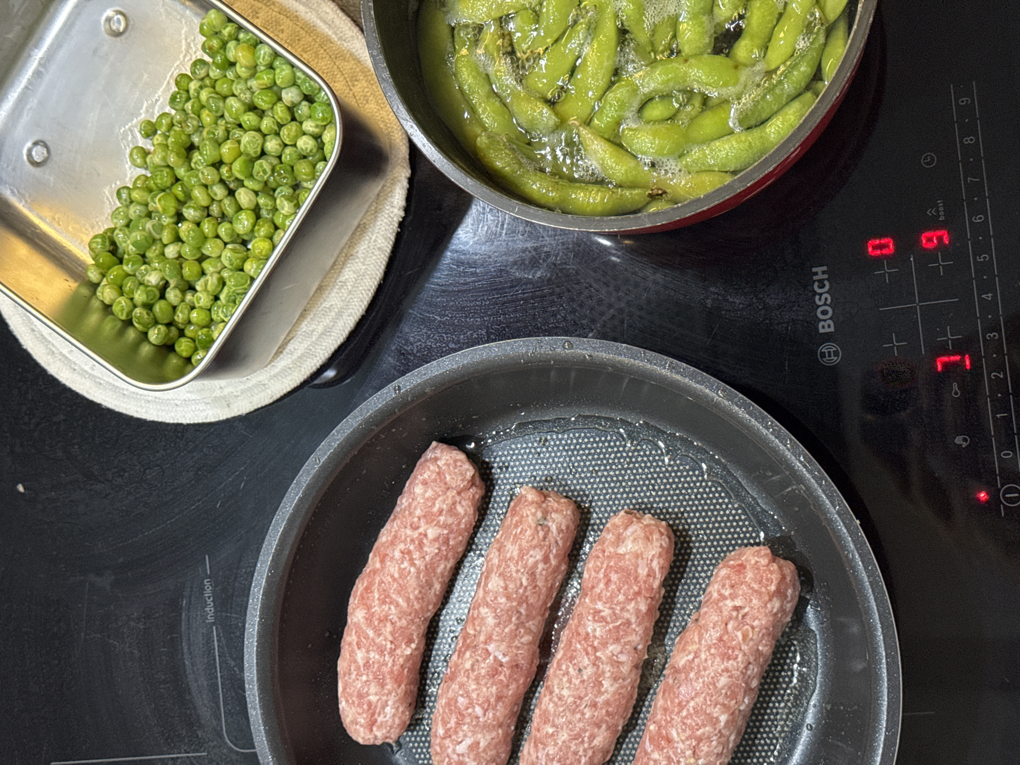 An overhead shot captures a cooking scene on a black Bosch induction cooktop in what is likely a kitchen in Bac Nha Trang Ward, Vietnam.
The image features various food items being prepared. In the upper left, a rectangular stainless steel tray holds a pile of bright green peas, possibly recently drained or shelled. To its right, a round pot contains numerous green edamame pods bubbling in hot water, suggesting they are being boiled. Below these, a frying pan holds four raw, pinkish-beige sausages, arranged longitudinally, resting in a small amount of oil, ready for or in the early stages of cooking.
The induction cooktop shows several digital readouts: a "0" is visible above a "+" button, a "9" with a "boost" function engaged is displayed above another "+", and a "7" is shown in the bottom right, all indicating active cooking zones and heat settings. The brand name "BOSCH" is clearly visible on the cooktop, along with the word "Induction" and numerical settings from "0" to "9". A small red light is illuminated near a power symbol, indicating the hob is on or a zone is hot. The overall scene depicts a meal being actively prepared.