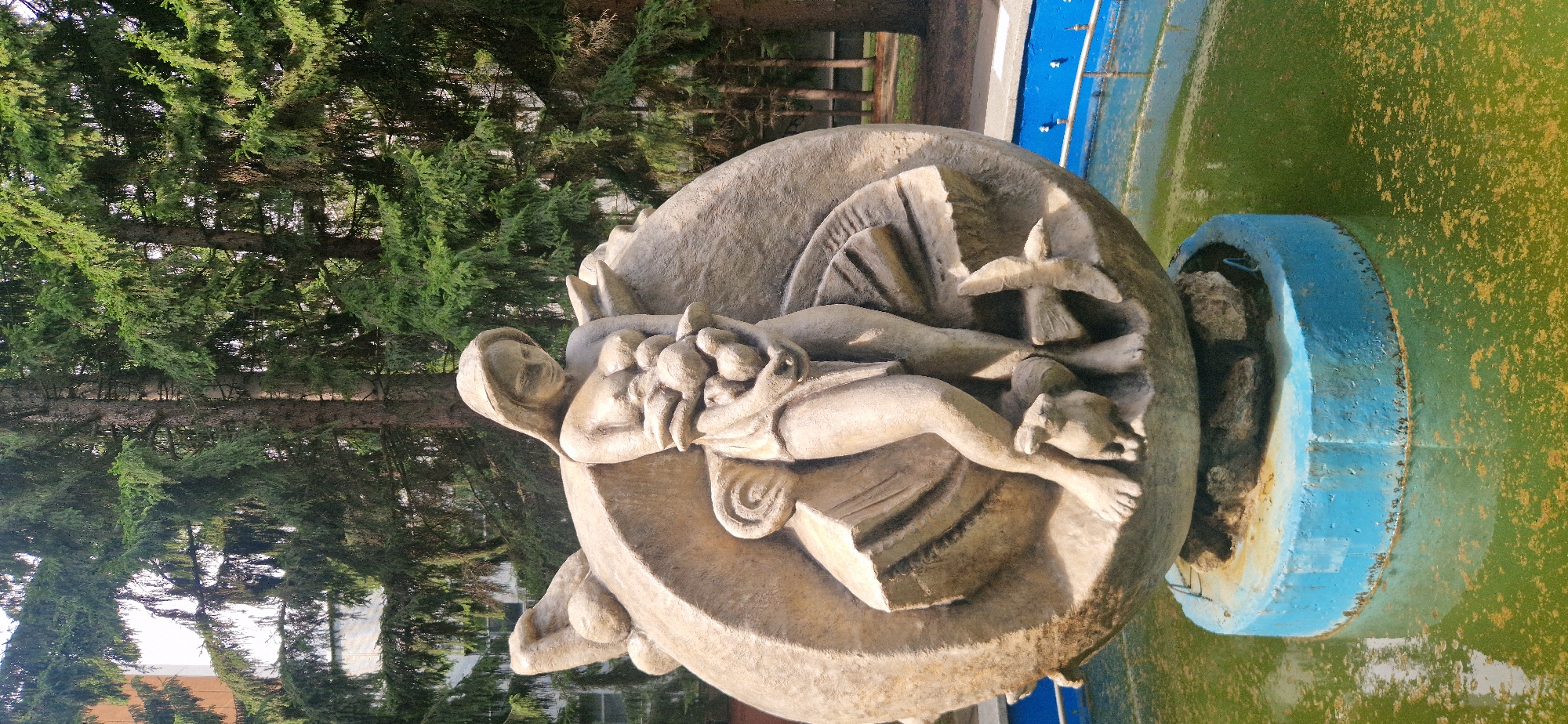 A light-colored stone fountain sculpture depicts a woman holding a cluster of objects, possibly fruit, with two birds and a small animal at her feet. The sculpture is mounted on a blue circular base within a green, somewhat murky pool of water. The background features tall evergreen trees and a light-colored building. This sculpture is located in the city of Krasnoobsk, Russia.