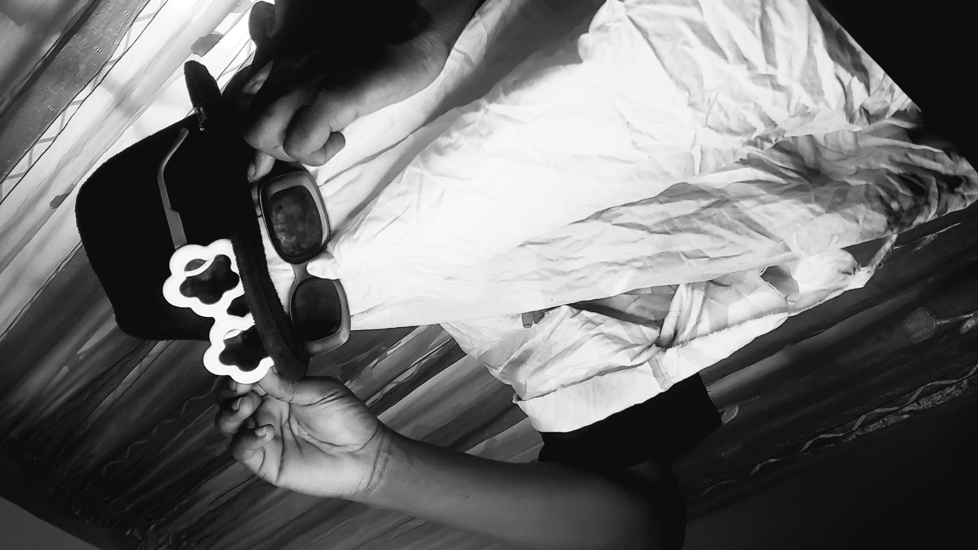 A person, partially obscured by a white cloth and wearing a black hat and sunglasses, adjusts their sunglasses. The person's hands are visible on either side of their face, touching the sunglasses and the hat. The background consists of vertically draped curtains. The scene is captured in black and white, with dramatic lighting that casts shadows and highlights the textures of the fabric and the person's skin. The image does not provide any information about the location in Narayi, Nigeria.
