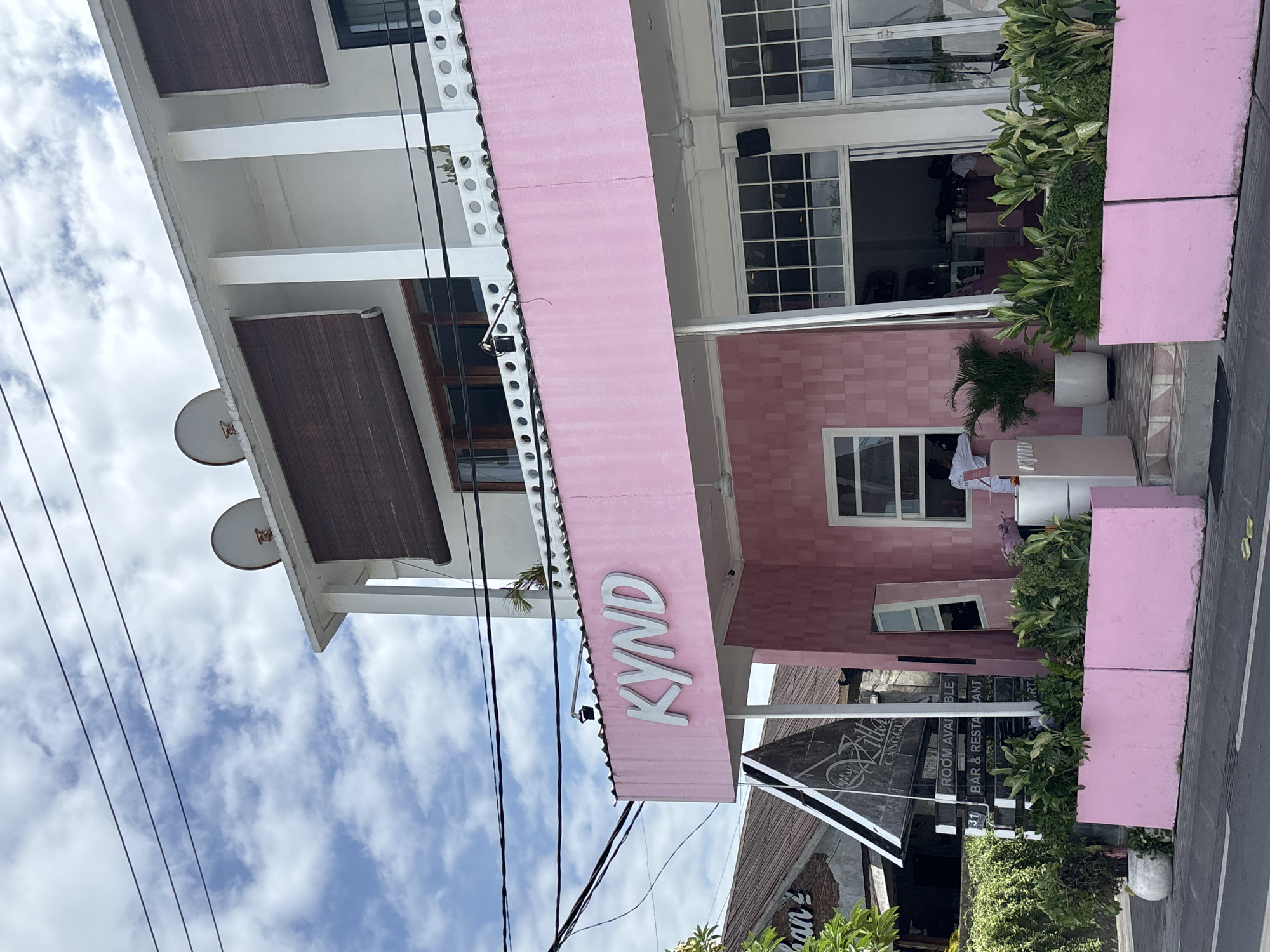 The image shows the exterior of a building in North Kuta, Indonesia. The main facade is pink with the word "KYND" in white capital letters on an overhanging awning. The building has multiple stories, with white architectural elements and dark brown blinds. There are windows with white frames. Plants in white pots are arranged along the entrance. Power lines are visible against a blue sky with clouds. A sign reading "My Killa Canggu" and "ROOM AVAILABLE BAR & RESTAURANT" is visible near the entrance. People are visible through the windows.